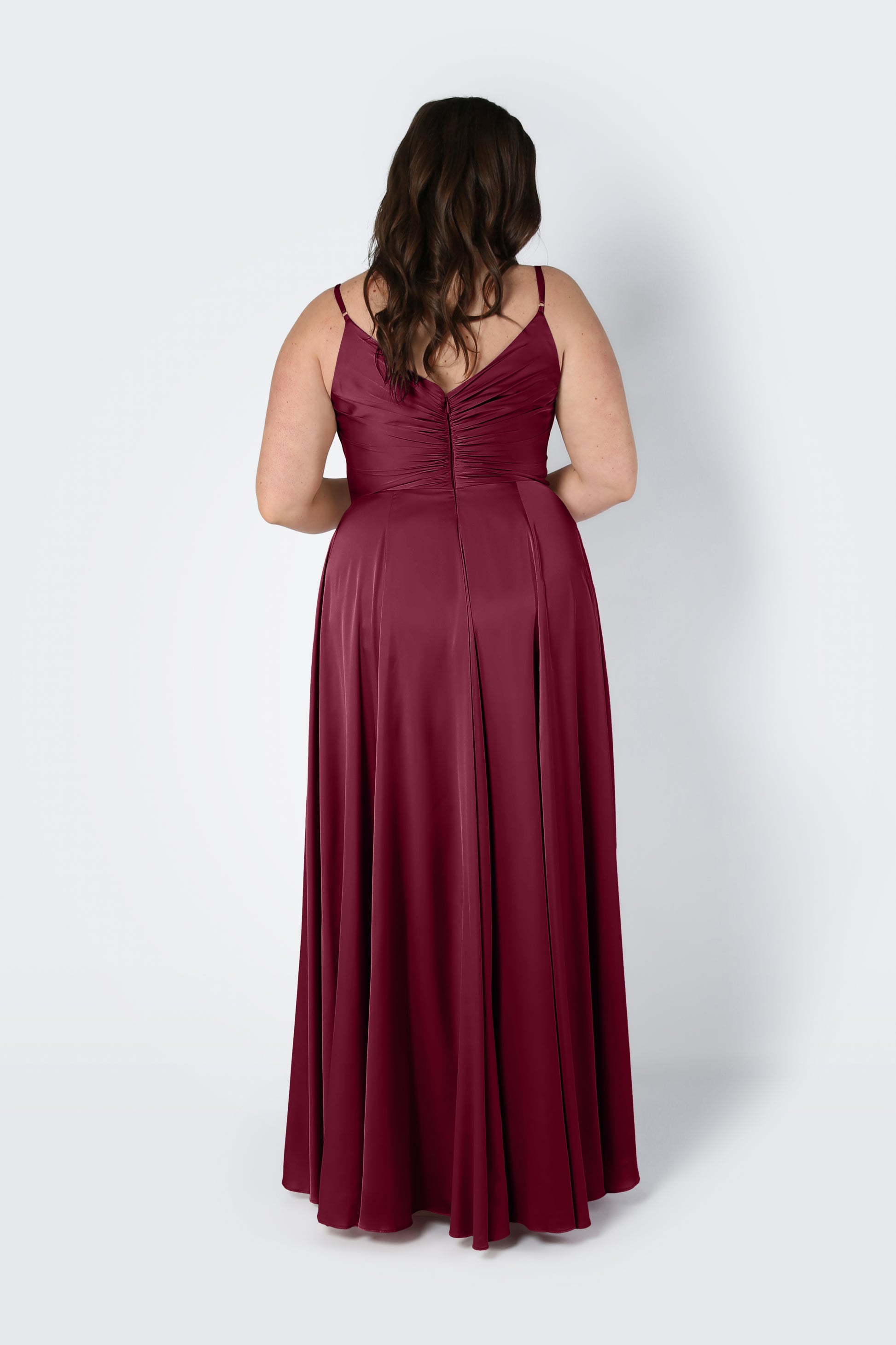 A woman with wavy brown hair stands with her back to the camera against a light background, wearing the TH&TH Isla Bridesmaid Dress in Merlot Satin—a sleeveless, floor-length dress with spaghetti straps and a pleated bodice.