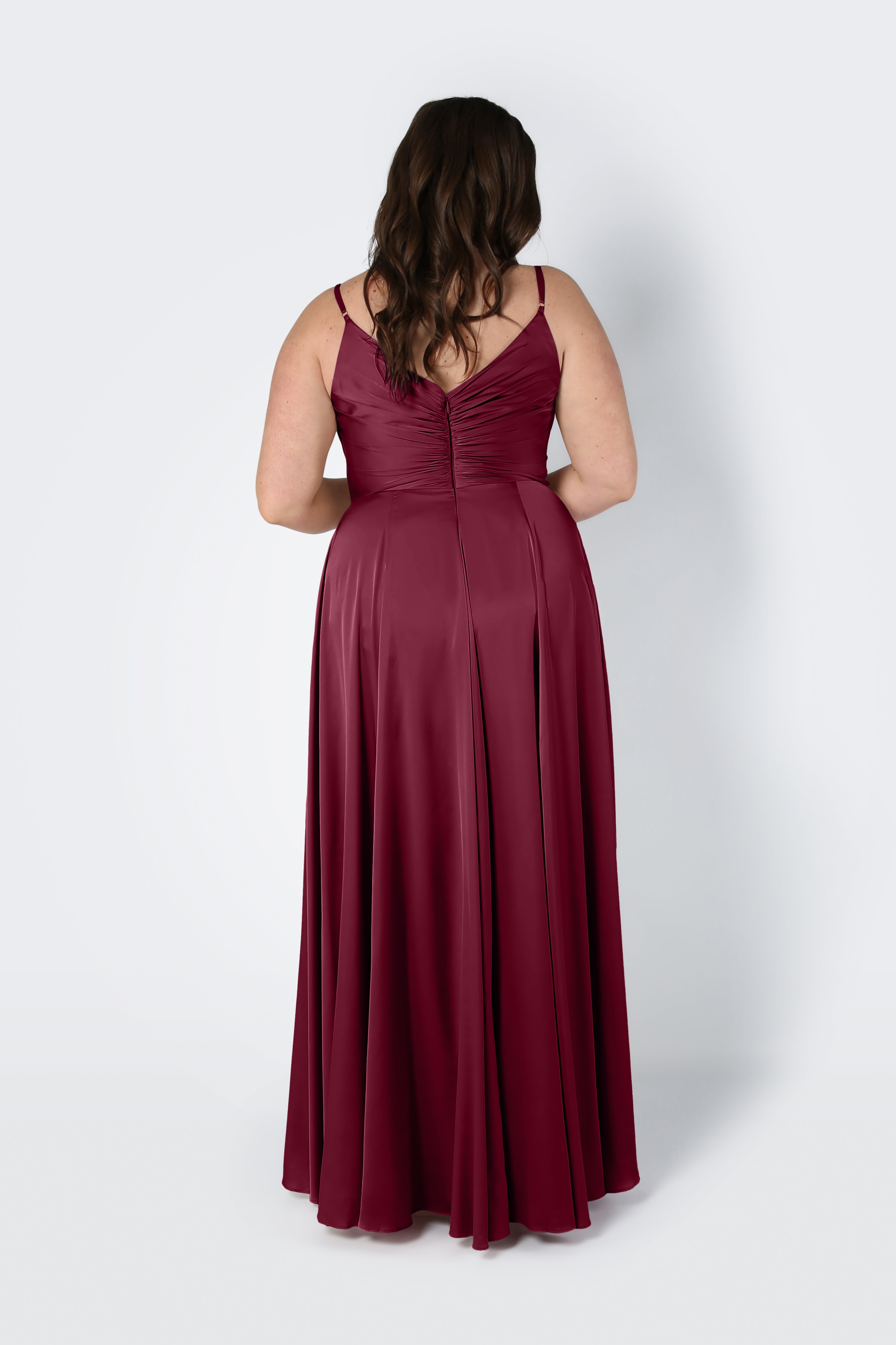 A woman with wavy brown hair stands with her back to the camera against a light background, wearing the TH&TH Isla Bridesmaid Dress in Merlot Satin—a sleeveless, floor-length dress with spaghetti straps and a pleated bodice.