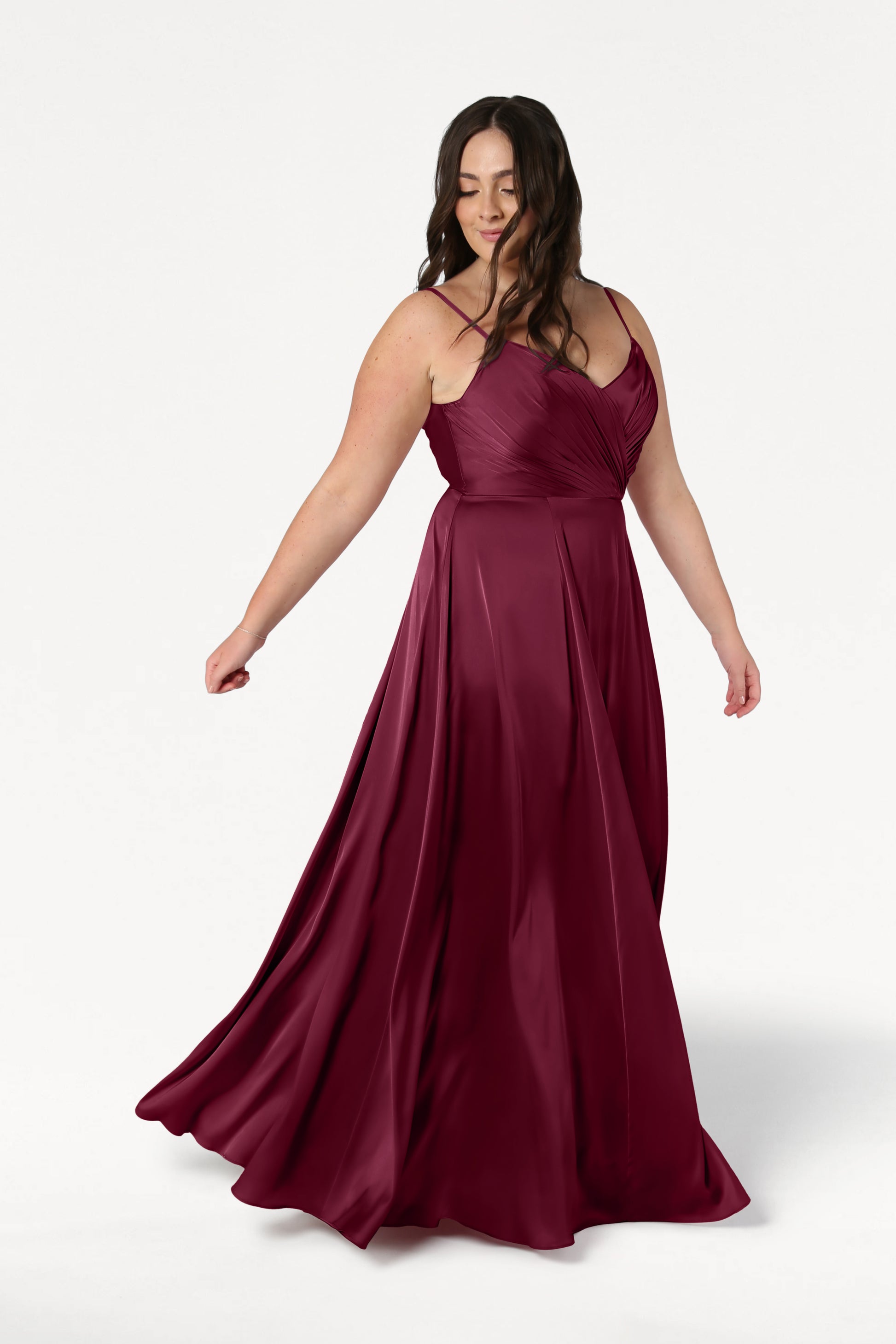 A woman in the TH&TH Isla Bridesmaid Dress in Merlot Satin, a sleeveless floor-length gown with a pleated bodice and flowing skirt, stands against a white background, looking down and gently holding her dress.