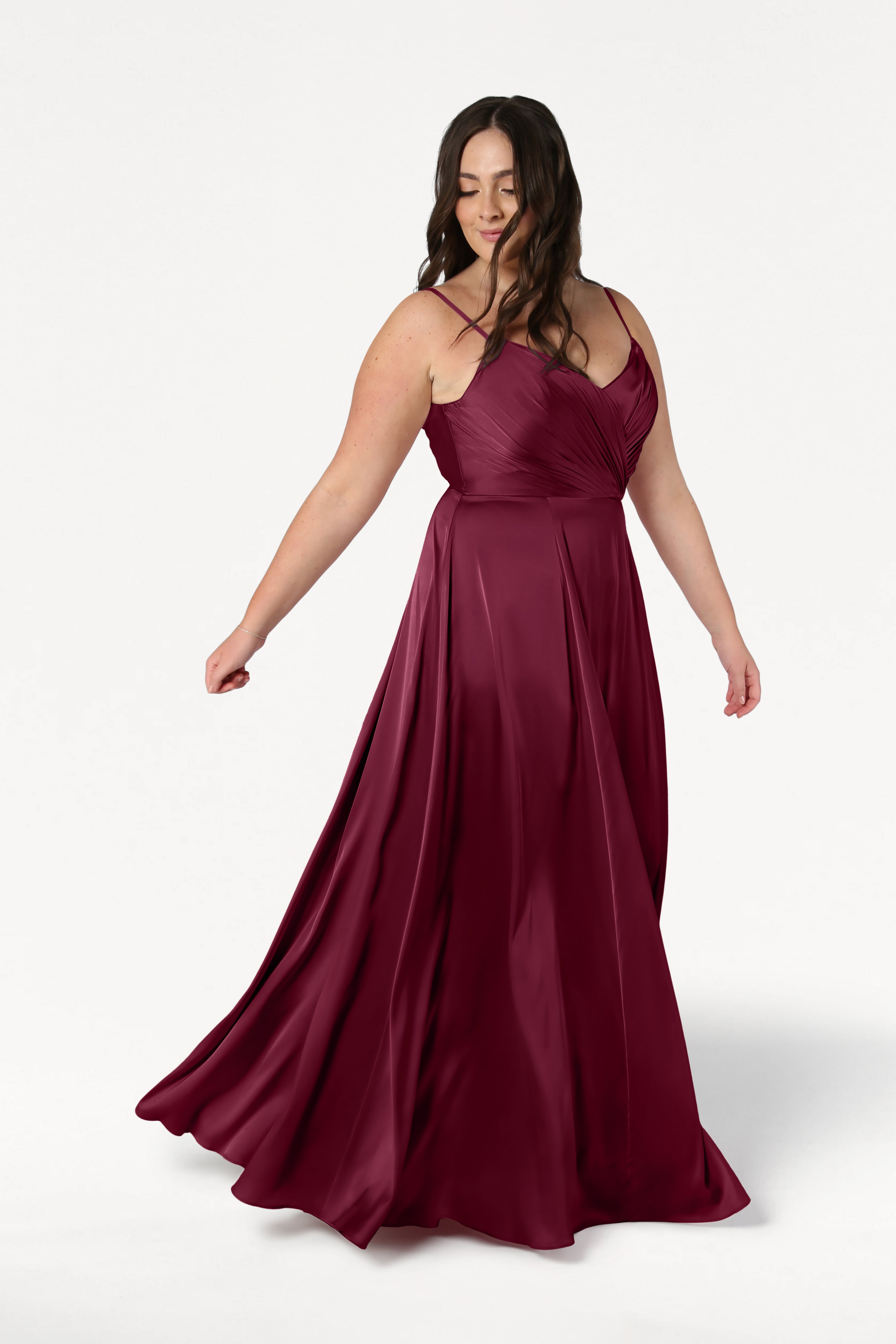 A woman in the TH&TH Isla Bridesmaid Dress in Merlot Satin, a sleeveless floor-length gown with a pleated bodice and flowing skirt, stands against a white background, looking down and gently holding her dress.