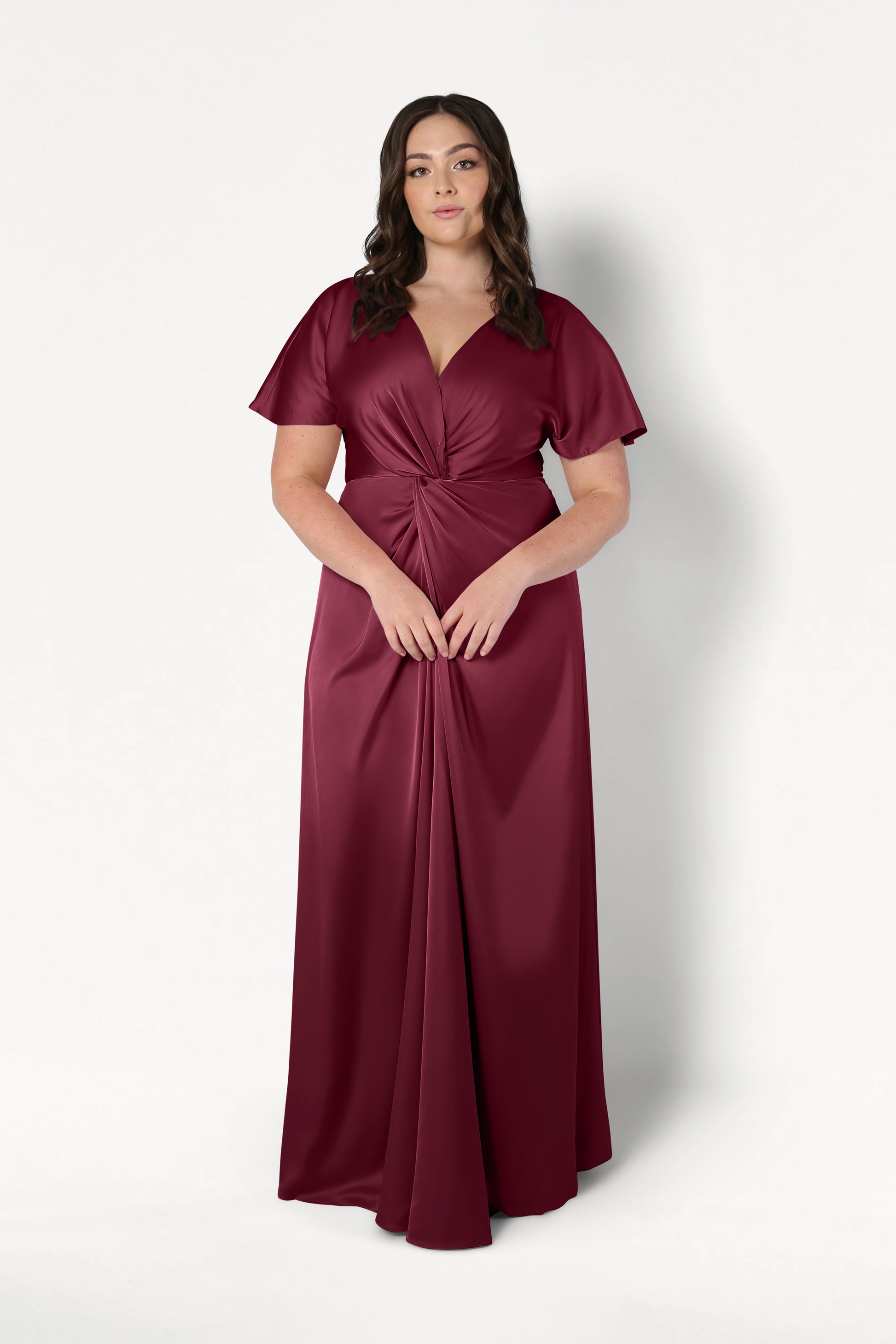 A woman wears the TH&TH Camilla Bridesmaid Dress in Merlot Satin, a floor-length gown with short sleeves, a V-neckline, and gathered knot waist detail, standing against a plain white background.