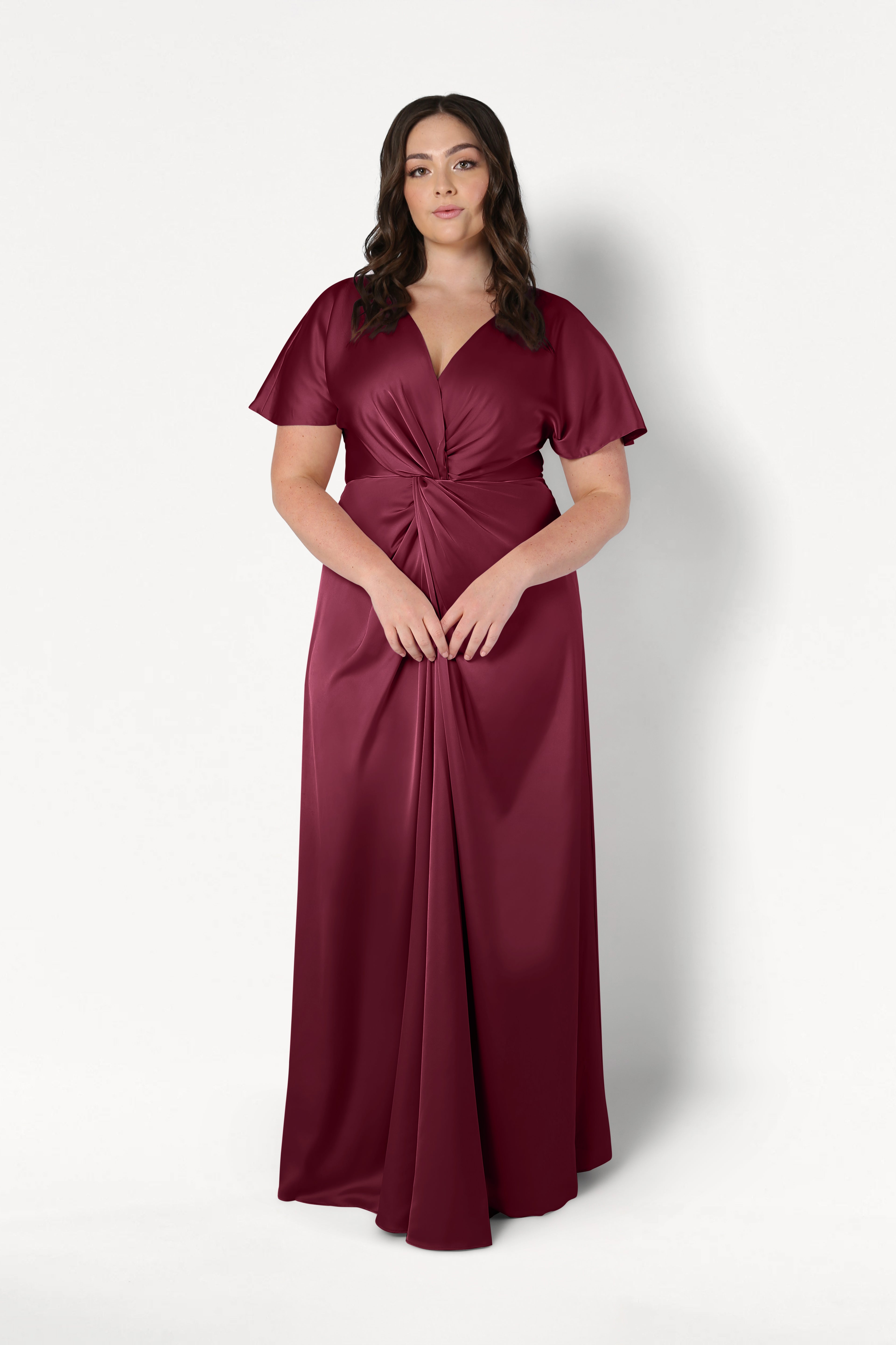 A woman wears the TH&TH Camilla Bridesmaid Dress in Merlot Satin, a floor-length gown with short sleeves, a V-neckline, and gathered knot waist detail, standing against a plain white background.