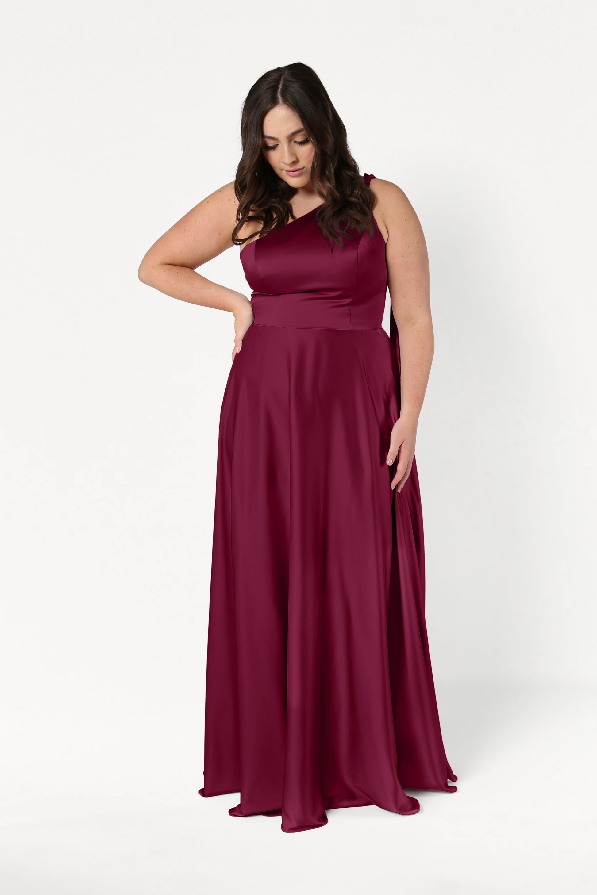 A woman models the TH&TH Lyra Bridesmaid Dress in Merlot Satin—a size-inclusive, floor-length gown—posing against a white background with one hand on her hip and the other by her side, gazing downward.