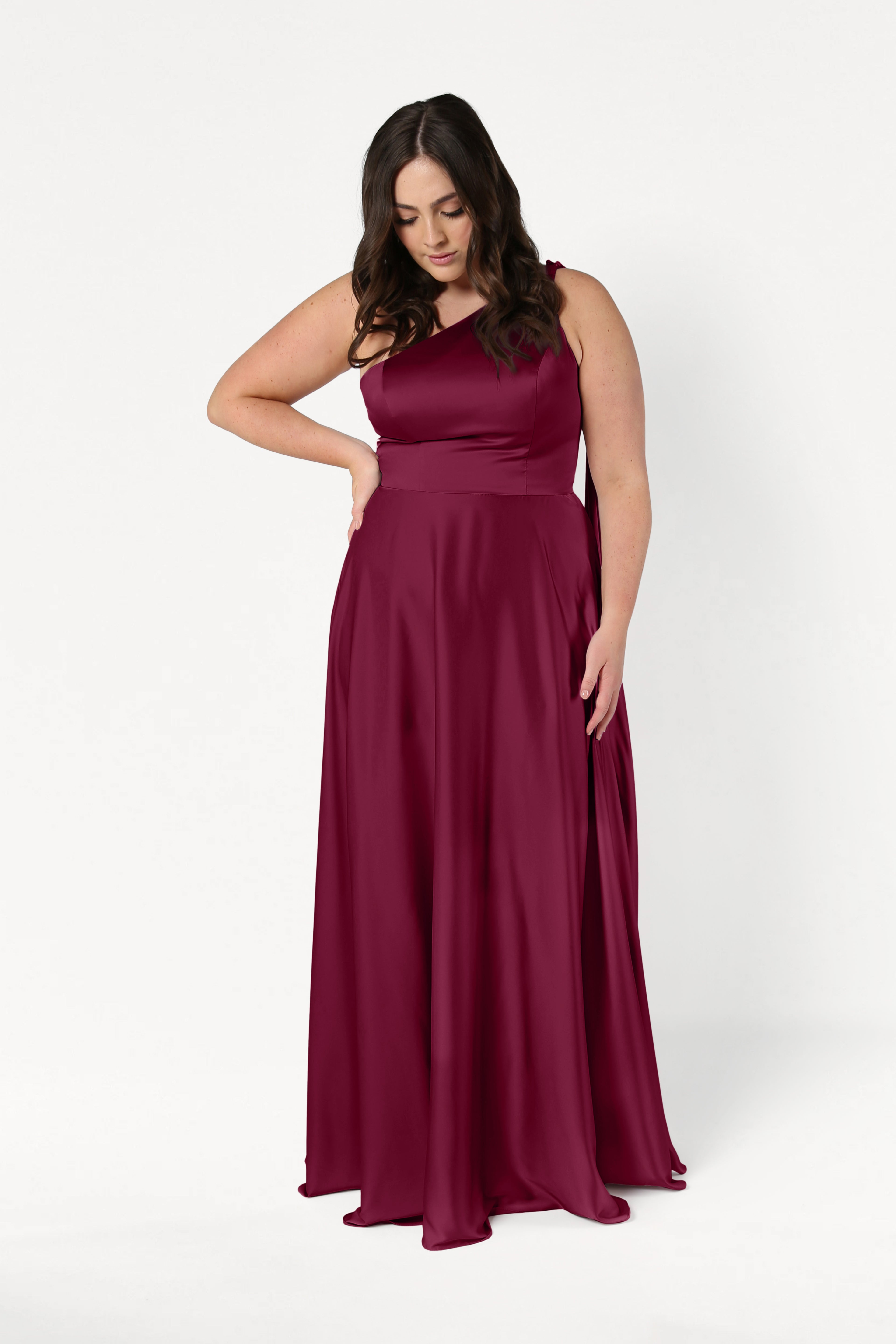 A woman models the TH&TH Lyra Bridesmaid Dress in Merlot Satin—a size-inclusive, floor-length gown—posing against a white background with one hand on her hip and the other by her side, gazing downward.