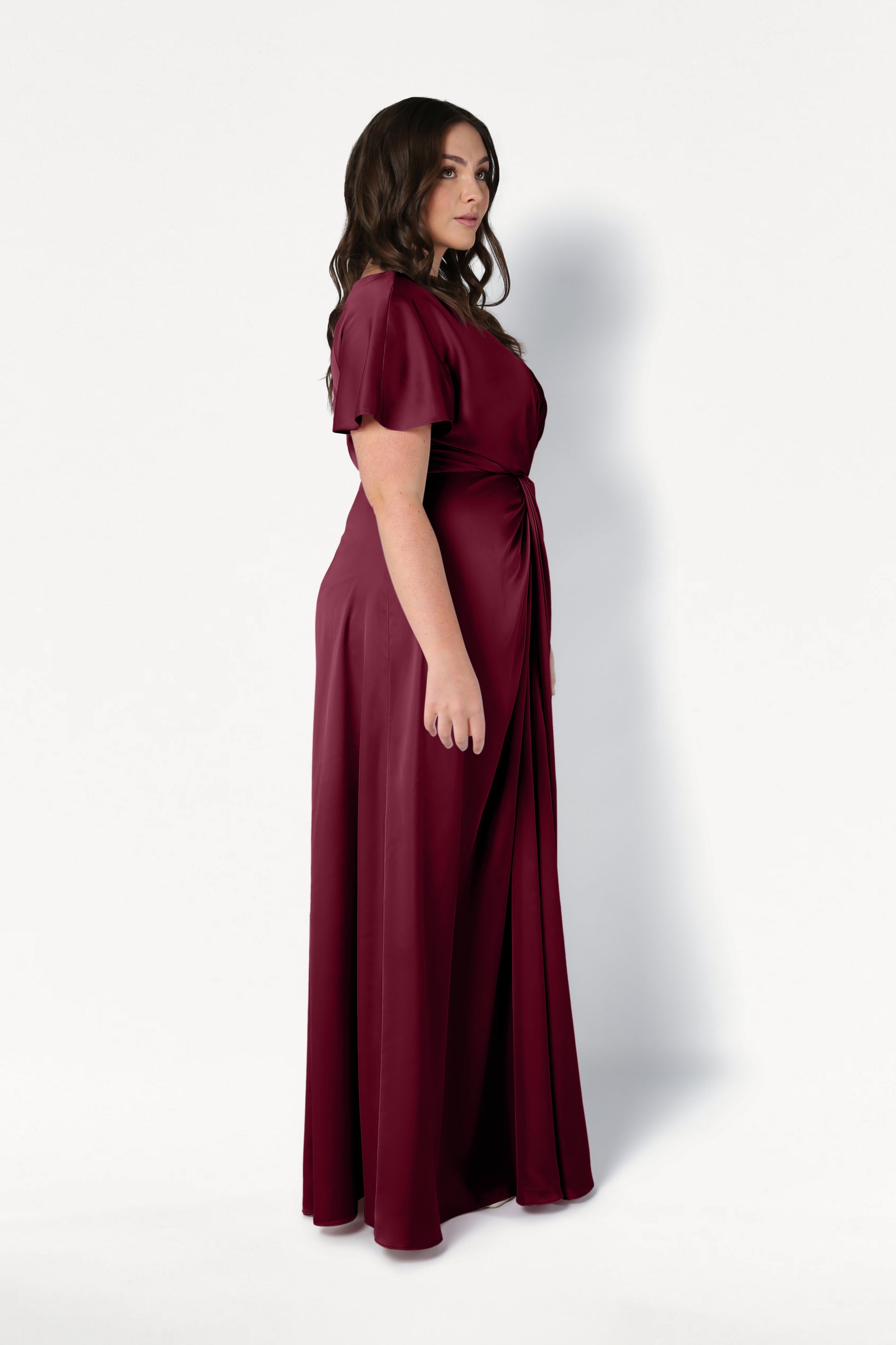 A woman with long dark hair stands in profile, wearing the TH&TH Camilla Bridesmaid Dress in Merlot Satin—a floor-length, short-sleeved gown with a gathered waist, showcased against a plain white background.