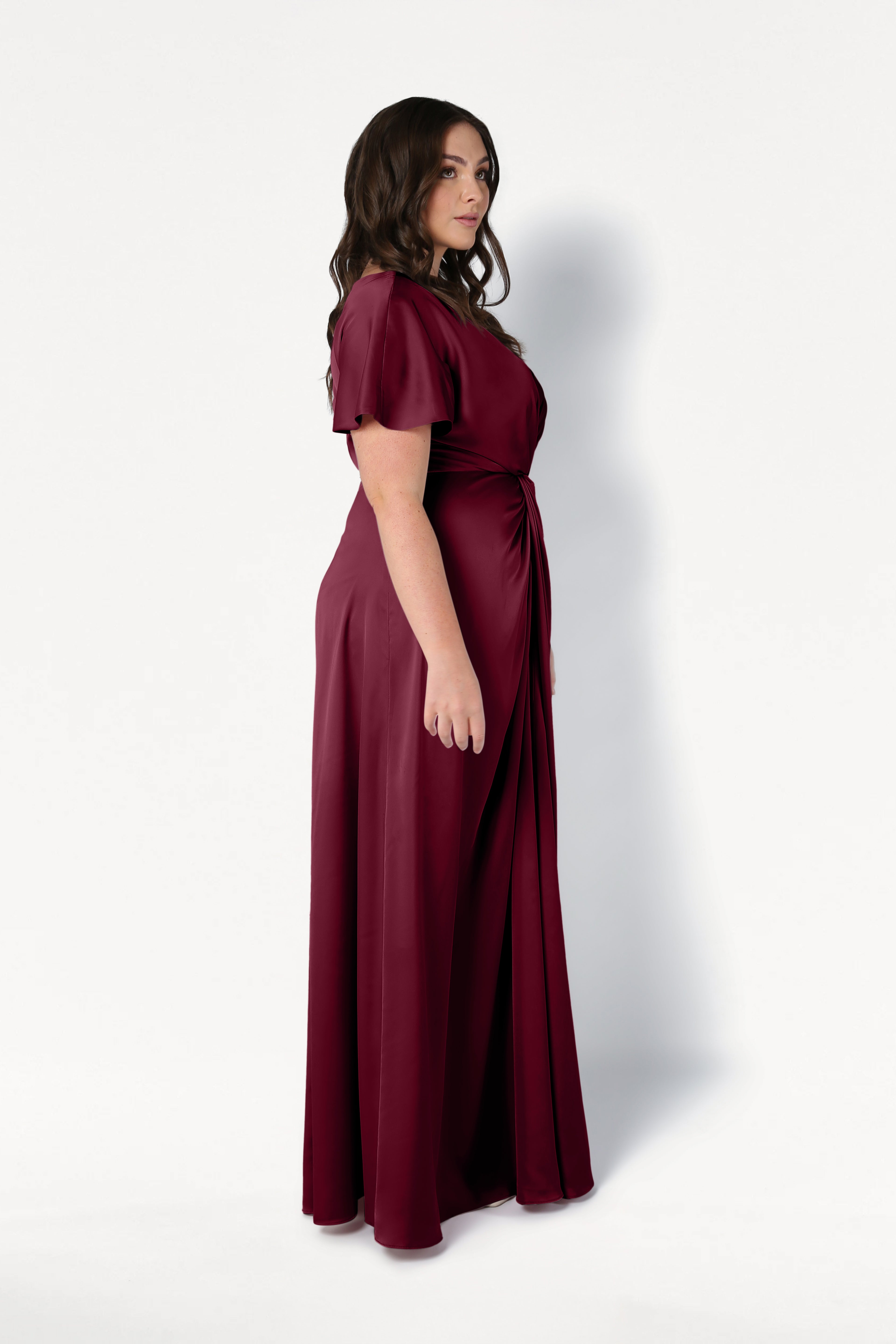 A woman with long dark hair stands in profile, wearing the TH&TH Camilla Bridesmaid Dress in Merlot Satin—a floor-length, short-sleeved gown with a gathered waist, showcased against a plain white background.
