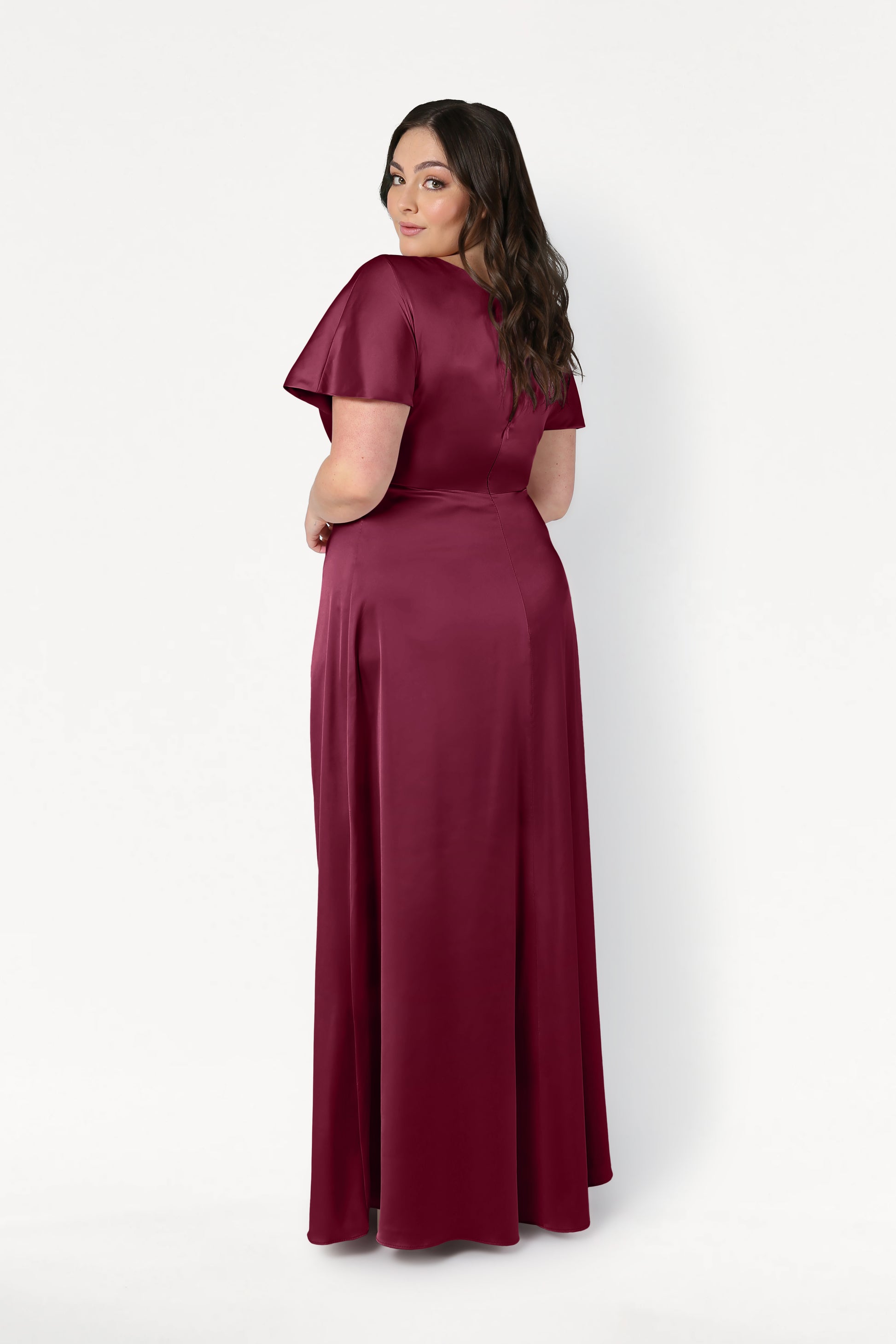 A woman with long dark hair looks over her shoulder, wearing the TH&TH Camilla Bridesmaid Dress in Merlot Satin—a modern, floor-length, short-sleeved gown—posing against a plain white background.