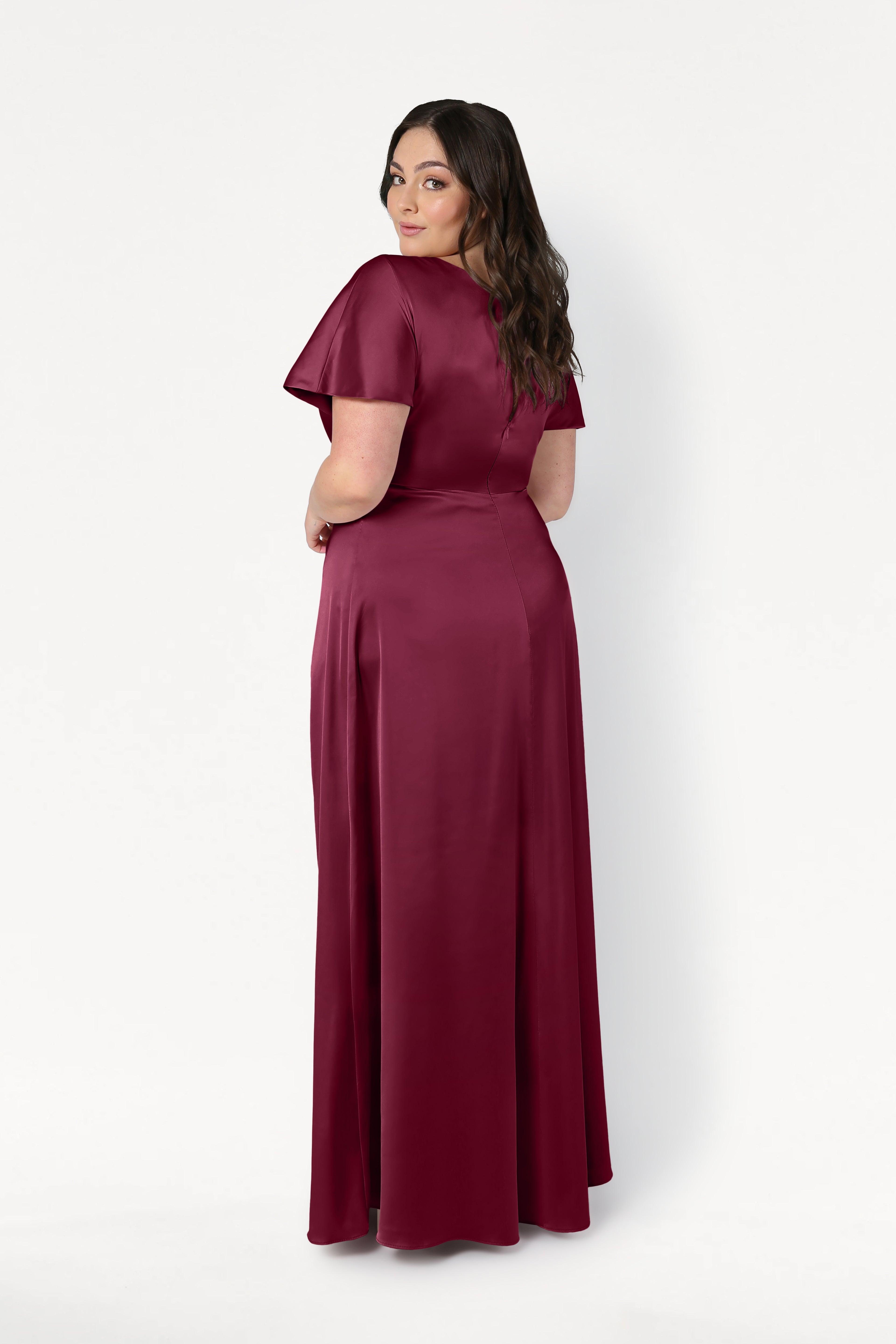 A woman with long dark hair looks over her shoulder, wearing the TH&TH Camilla Bridesmaid Dress in Merlot Satin—a modern, floor-length, short-sleeved gown—posing against a plain white background.
