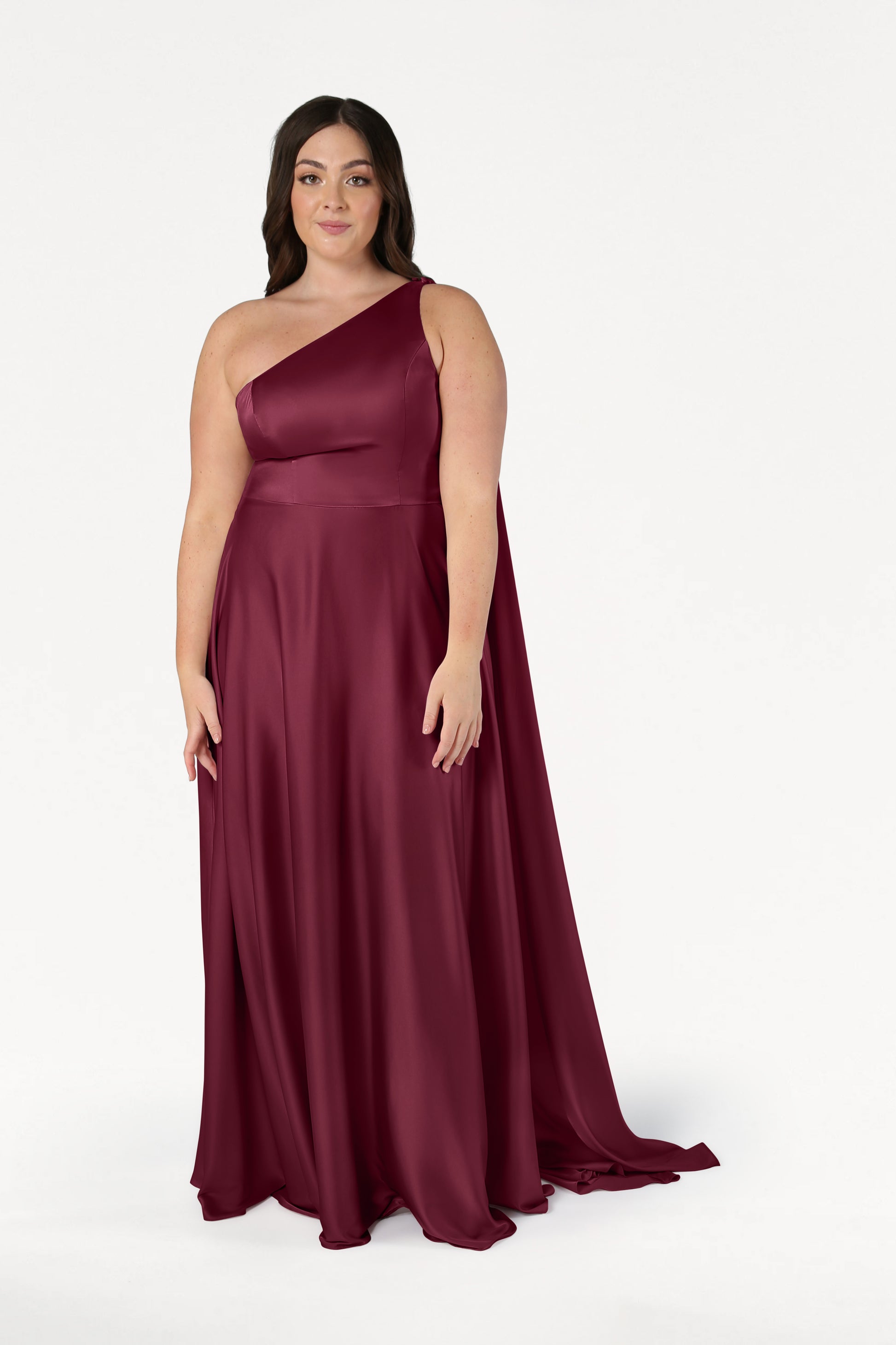 A woman wears the TH&TH Lyra Bridesmaid Dress in Merlot Satin, a size-inclusive, one-shoulder gown, standing against a plain white background.