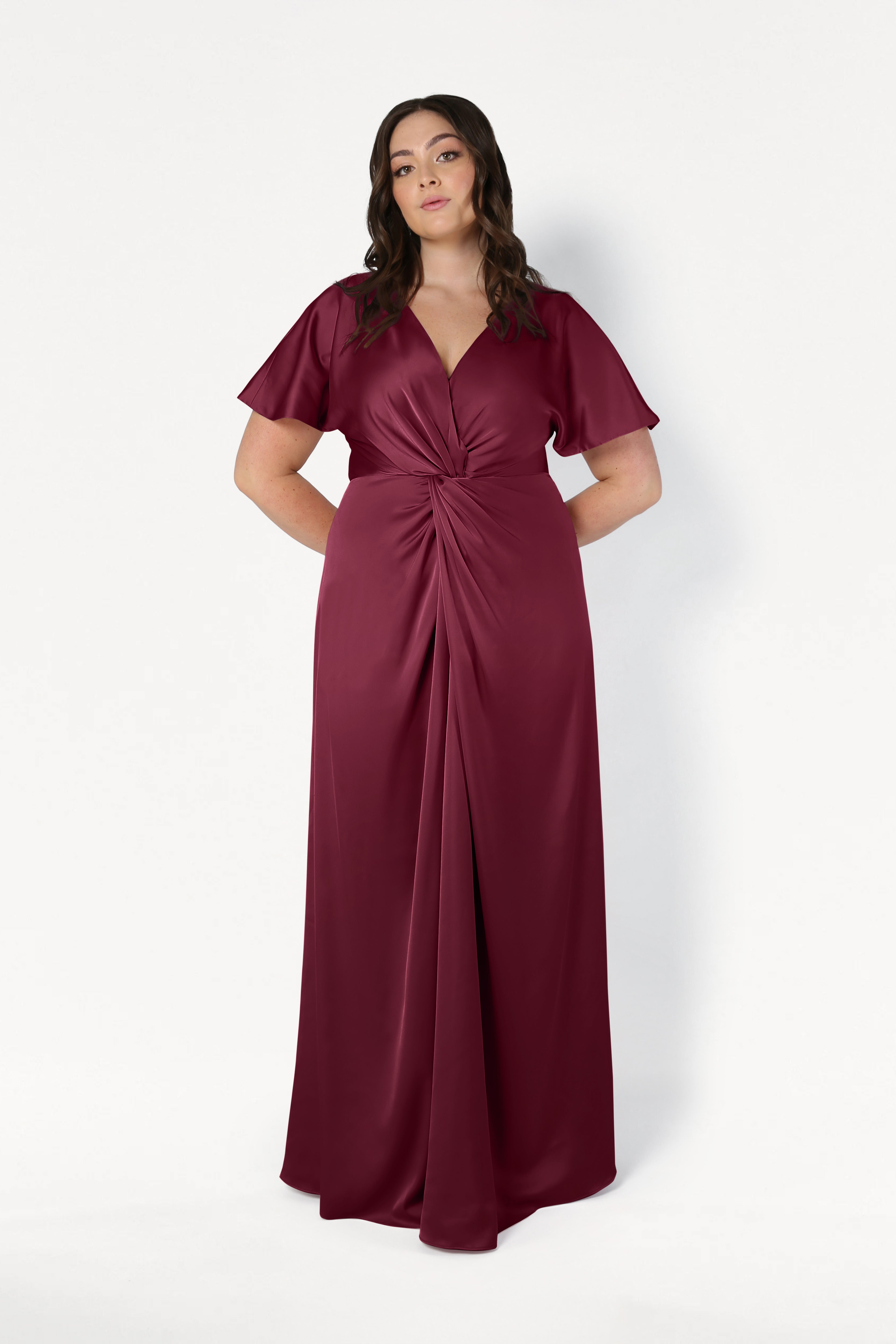 A woman stands against a plain white background, hands behind her back, wearing the TH&TH Camilla Bridesmaid Dress in Merlot Satin: a floor-length, short-sleeved gown with a gathered twist front detail.