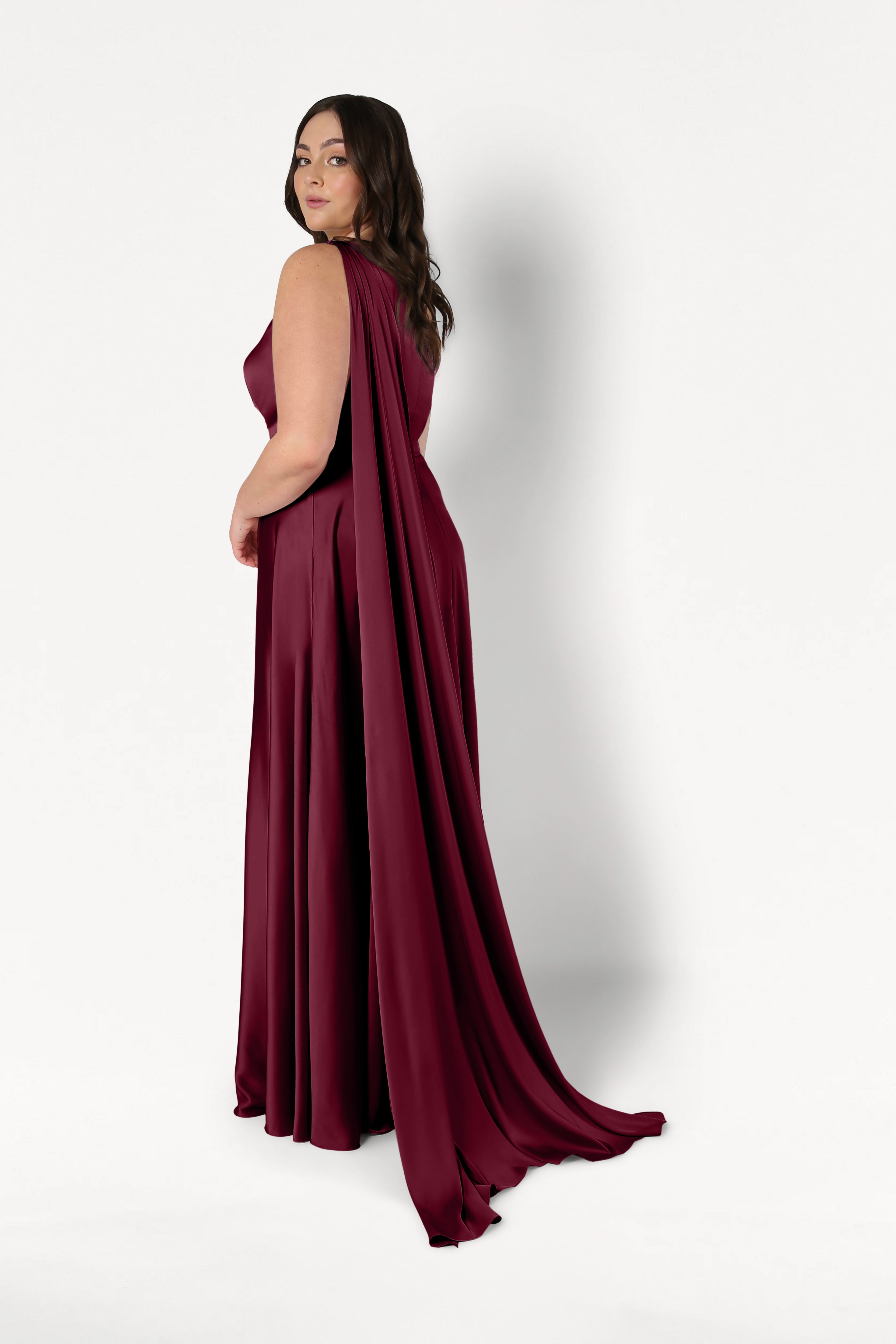 A woman poses in the Lyra Bridesmaid Dress in Merlot Satin by TH&TH, a size-inclusive, one-shoulder gown with a flowing cape, standing slightly turned against a plain white background.
