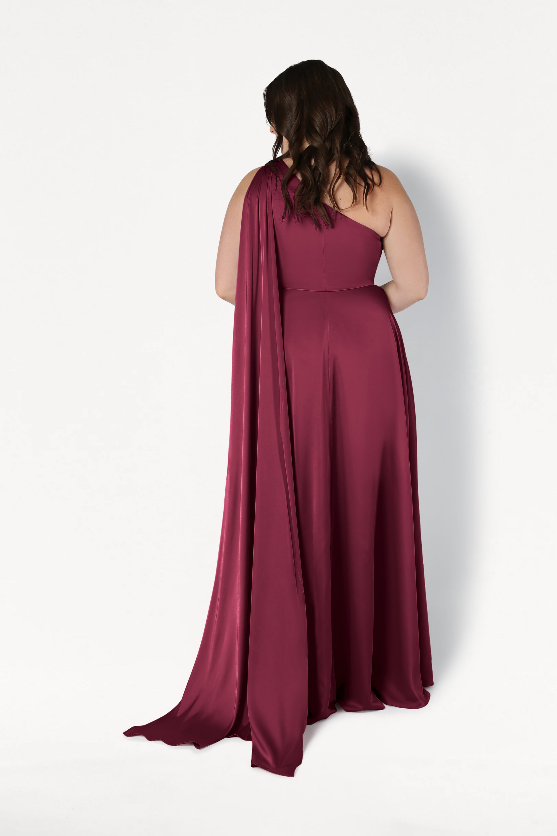 A woman with wavy brown hair faces away from the camera, modeling the TH&TH Lyra Bridesmaid Dress in Merlot Satin—a size-inclusive, floor-length one-shoulder gown with a flowing drape—against a plain white background.