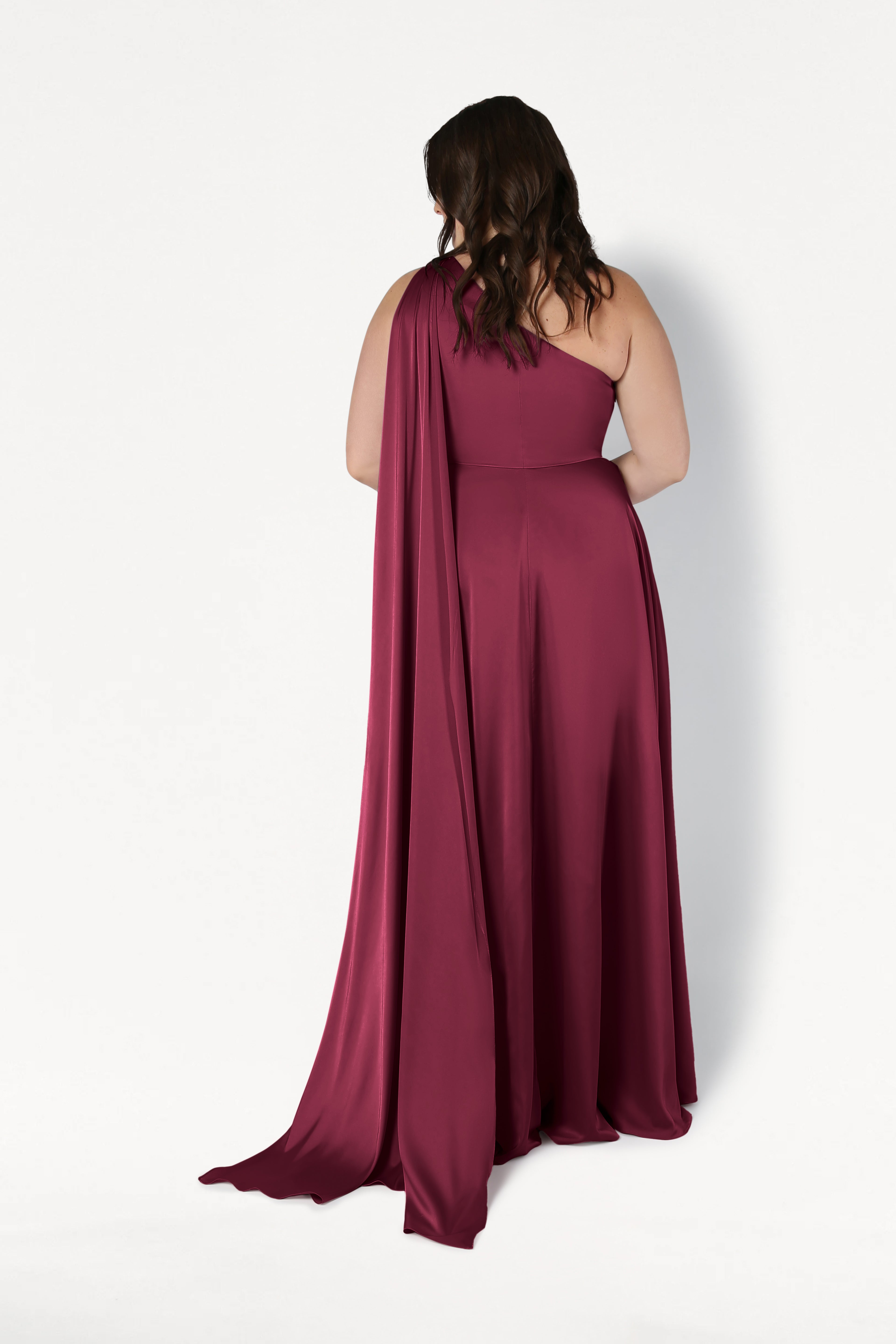A woman with wavy brown hair faces away from the camera, modeling the TH&TH Lyra Bridesmaid Dress in Merlot Satin—a size-inclusive, floor-length one-shoulder gown with a flowing drape—against a plain white background.