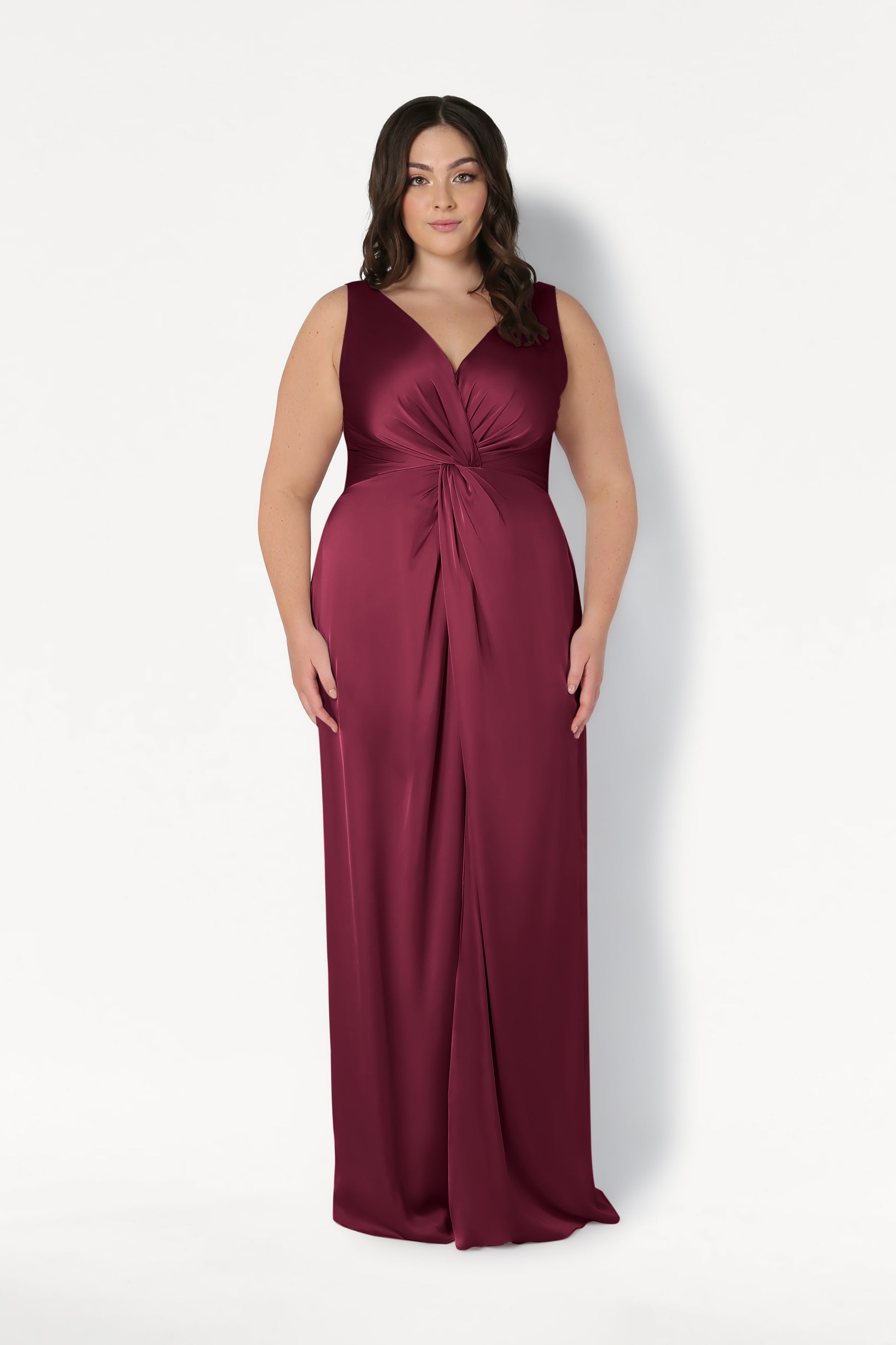 A woman with wavy dark hair wears the TH&TH Carmen Bridesmaid Dress in Merlot Satin, a sleeveless, floor-length gown with a V-neck and twist-knot waist detail, standing against a plain white background.