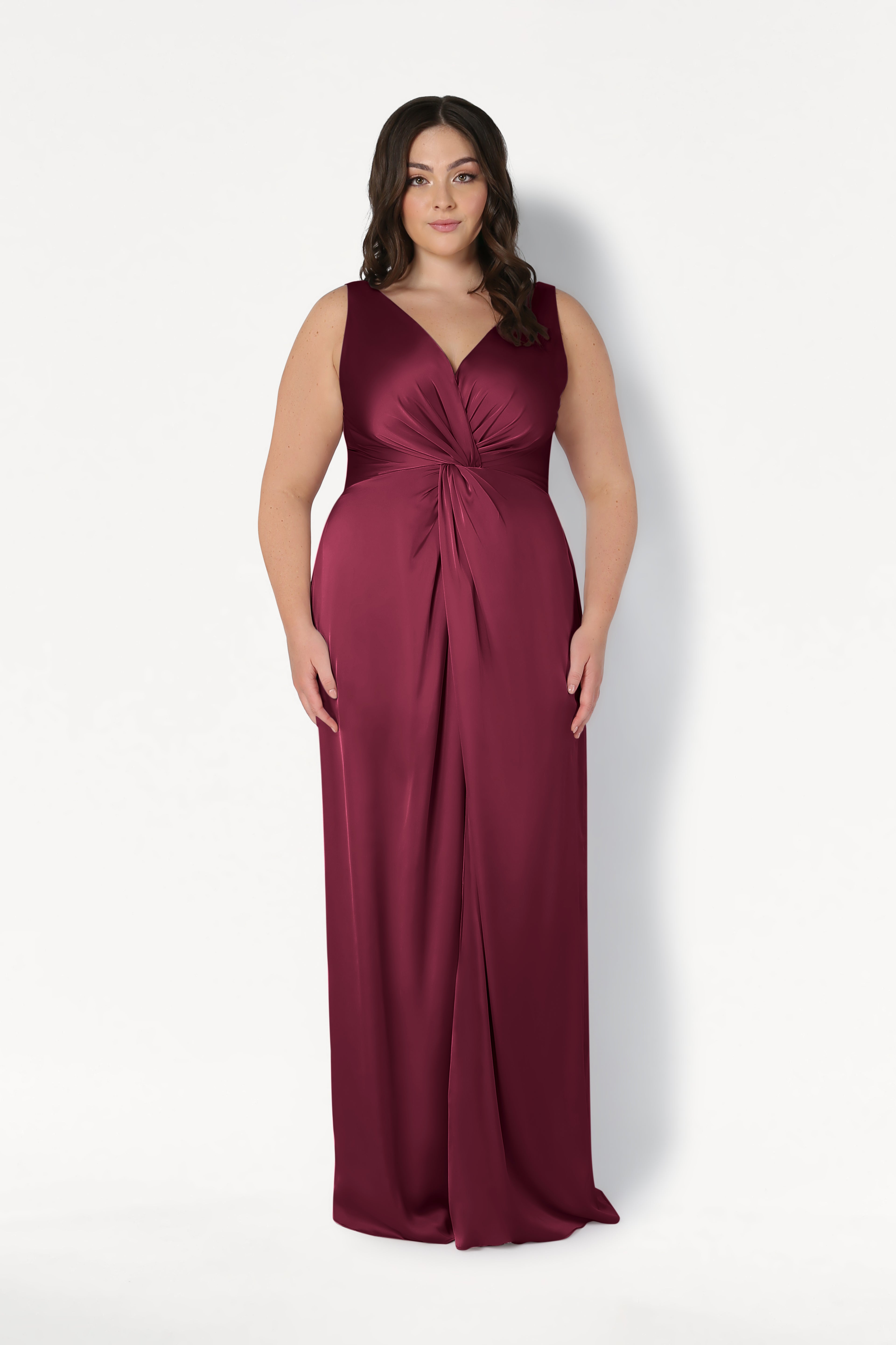 A woman with wavy dark hair wears the TH&TH Carmen Bridesmaid Dress in Merlot Satin, a sleeveless, floor-length gown with a V-neck and twist-knot waist detail, standing against a plain white background.