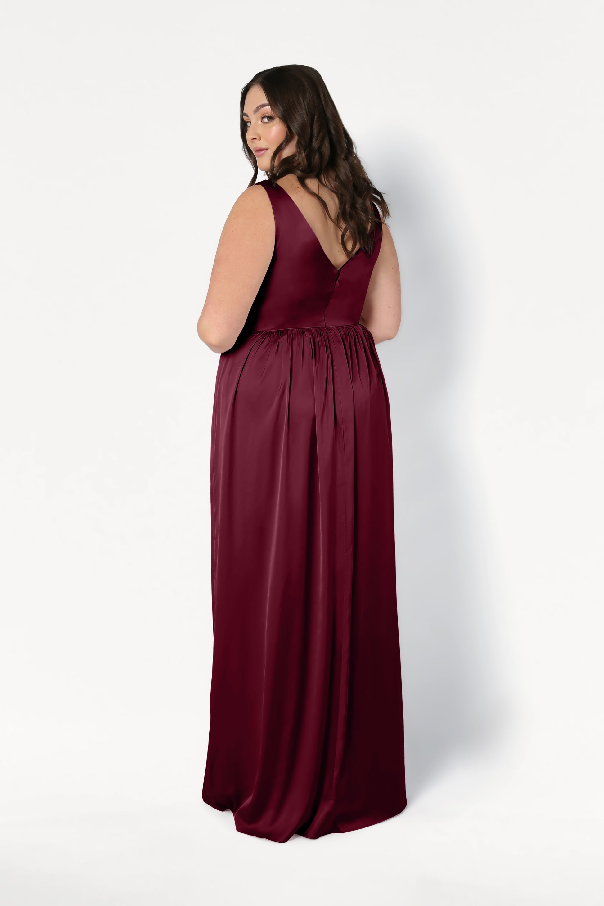 A woman models the TH&TH Carmen Bridesmaid Dress in Merlot Satin, a sleeveless, floor-length gown with a deep V-back and gathered waist, standing against a white background and looking over her shoulder with a gentle smile.