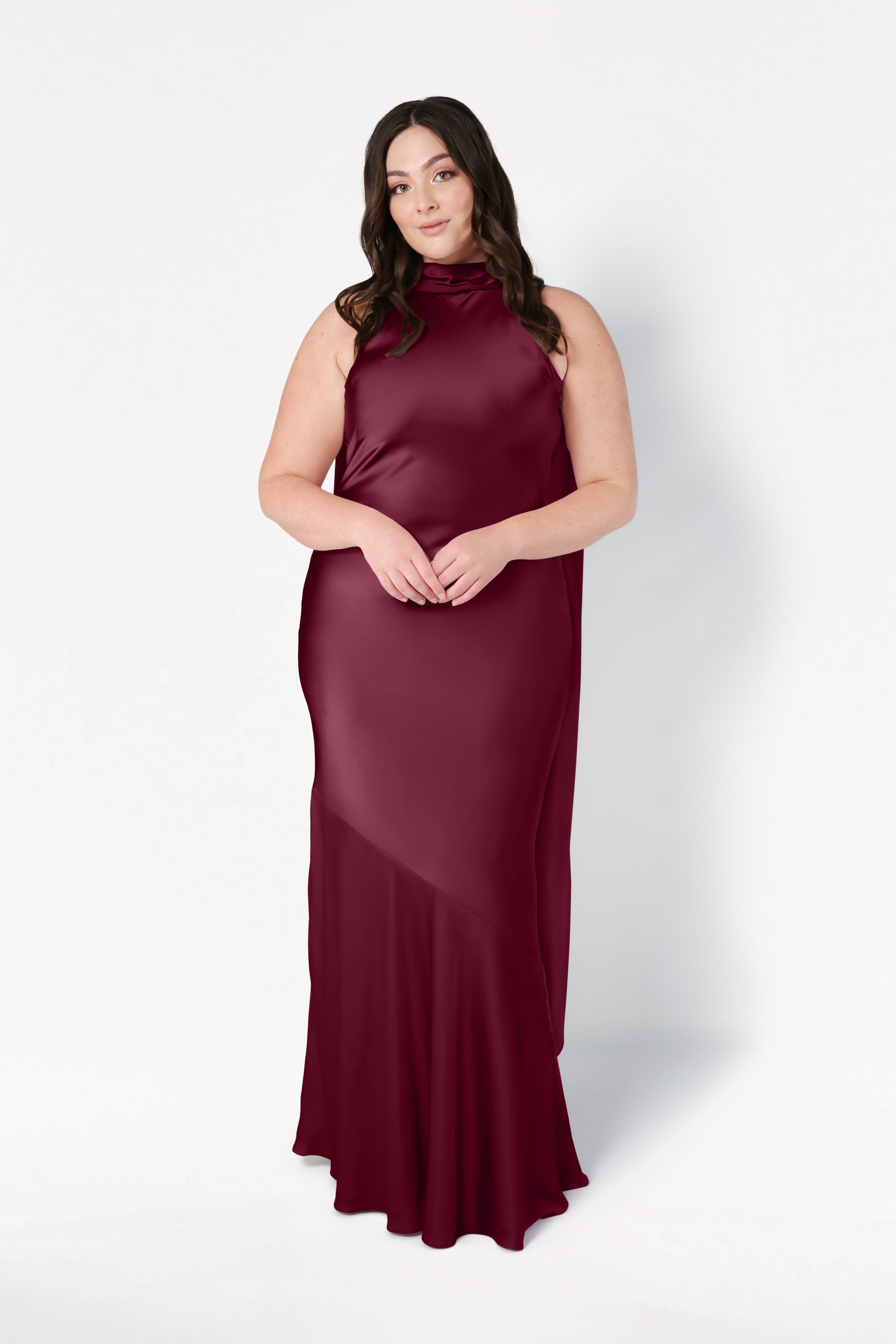 A woman with long brown hair styled in loose waves wears the TH&TH Mira Bridesmaid Dress in Merlot Satin, sleeveless and floor-length, standing against a plain white background.