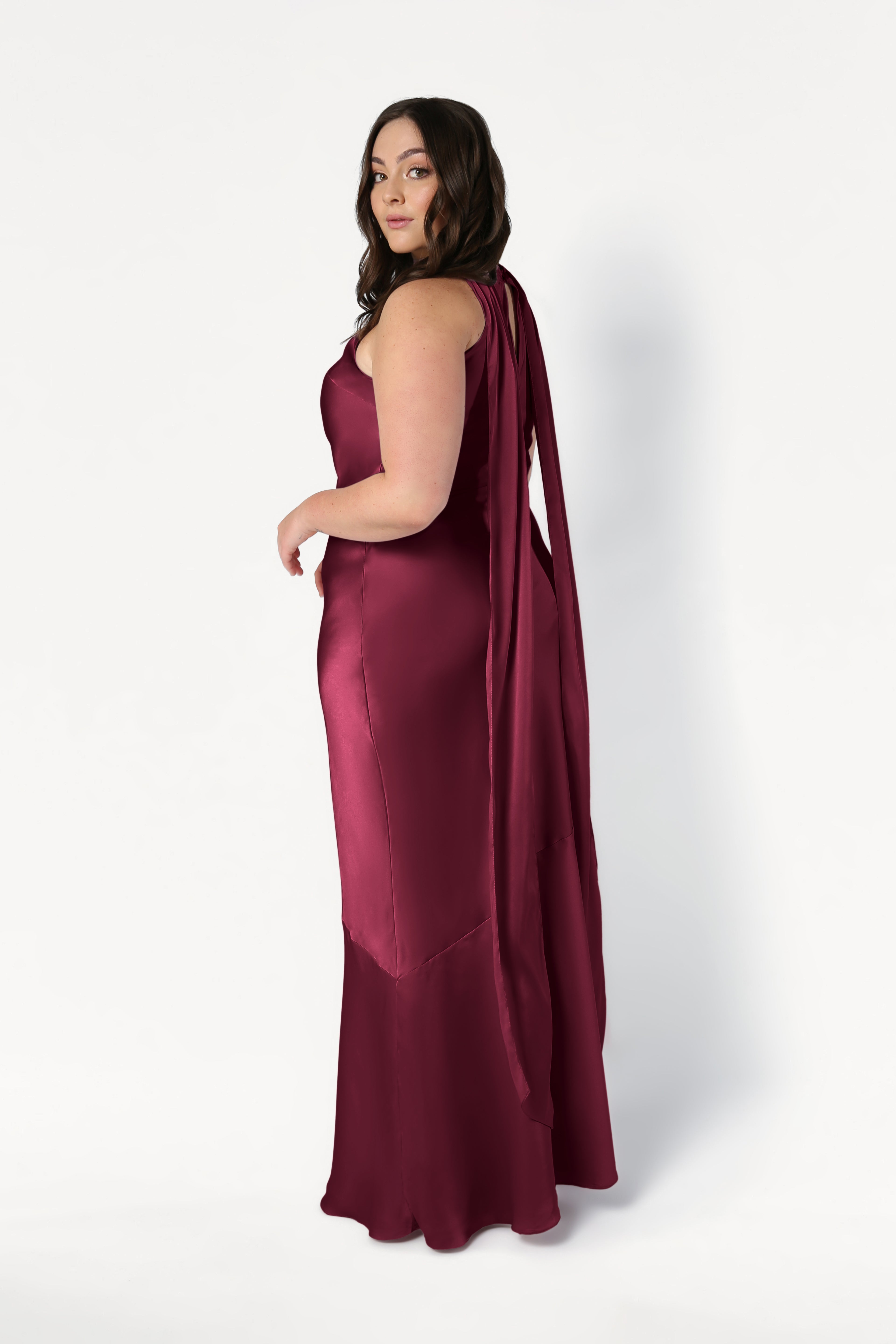 A woman with long brown hair poses against a white background in the TH&TH Mira Bridesmaid Dress in Merlot Satin, a sleeveless, floor-length gown featuring a draped back detail.