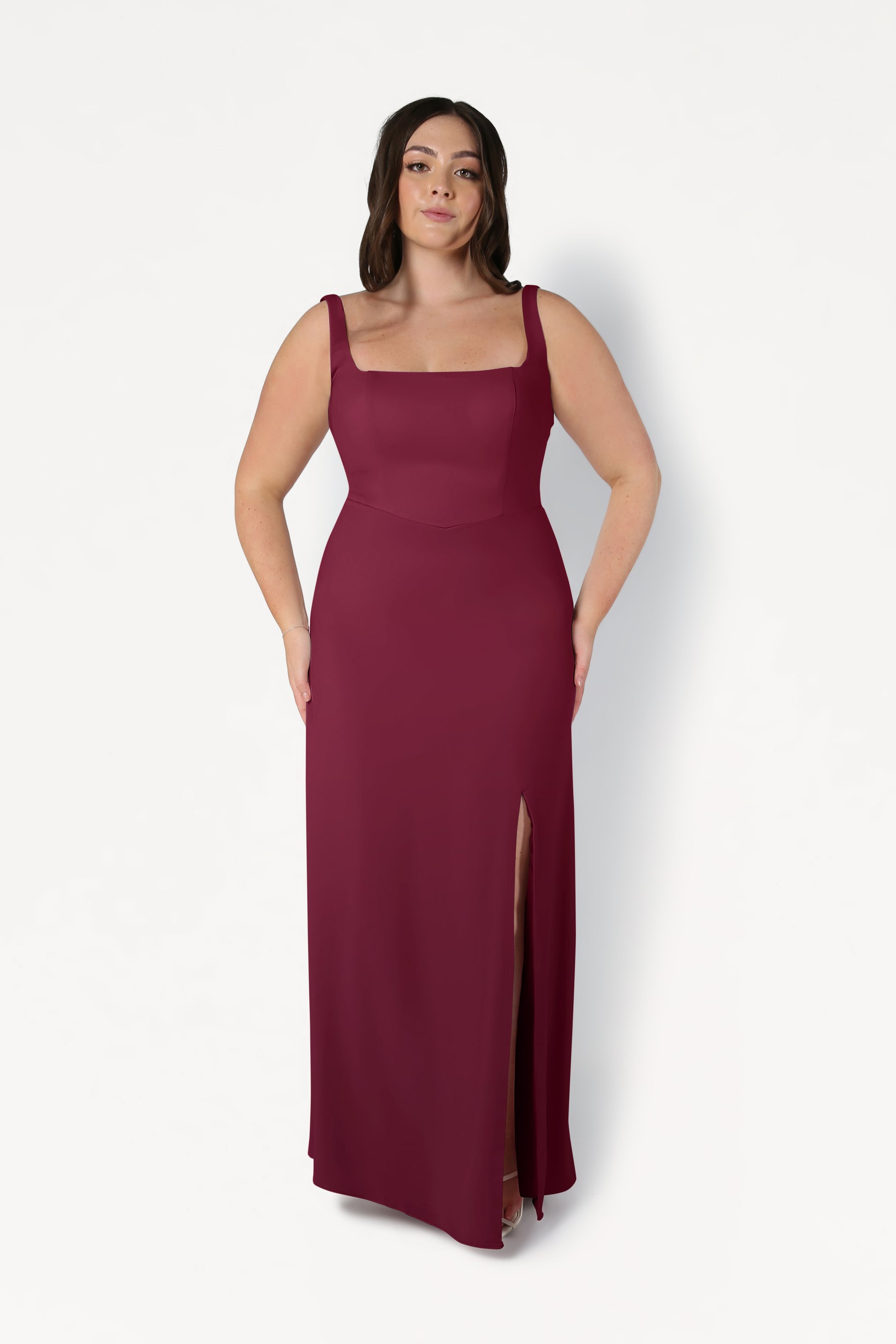 A woman models the TH&TH Helena Bridesmaid Dress in Merlot, featuring a sleeveless floor-length moss crepe design with a square neckline and thigh-high slit, standing against a plain white background.