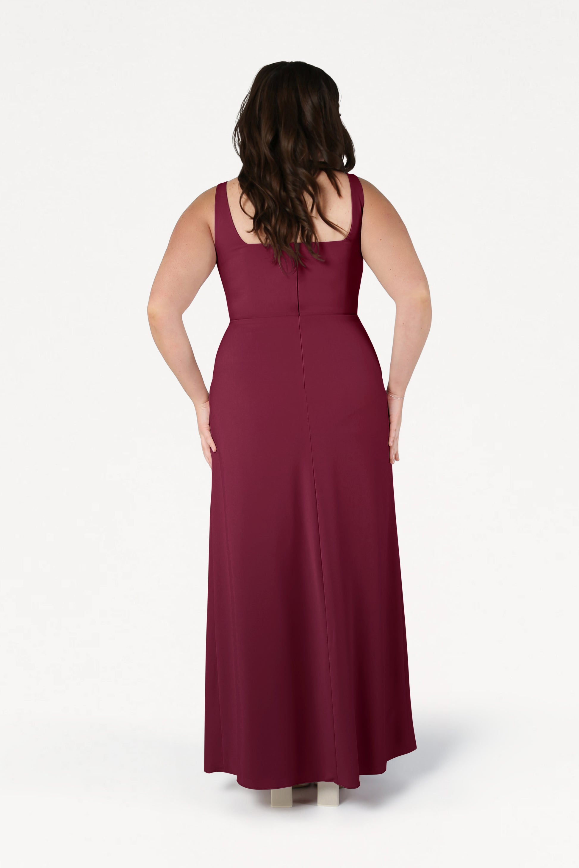 A woman with long, wavy brown hair stands with her back to the camera, modeling the TH&TH Helena Bridesmaid Dress in Merlot—a sleeveless, floor-length, size-inclusive gown—against a plain white background.