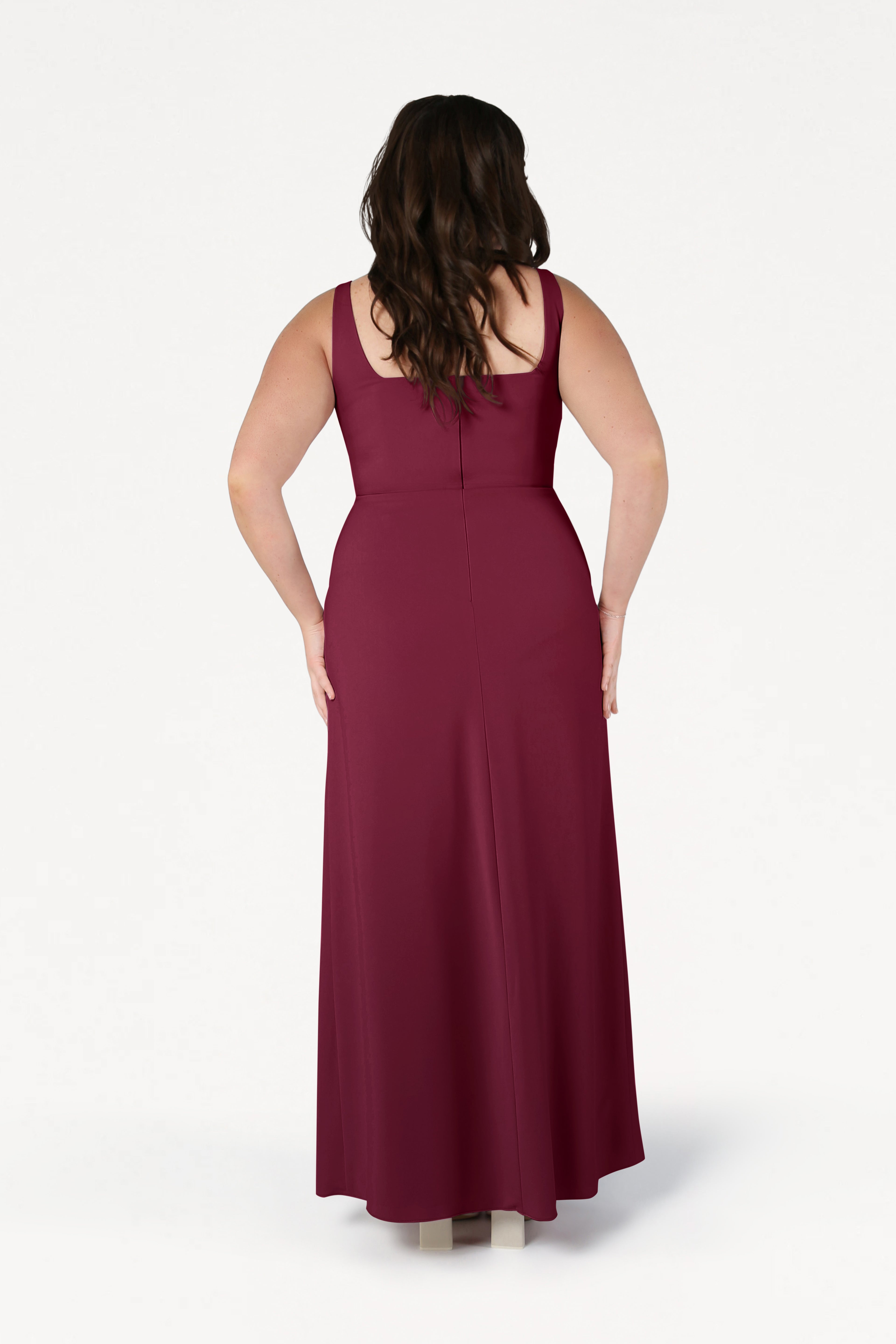 A woman with long, wavy brown hair stands with her back to the camera, modeling the TH&TH Helena Bridesmaid Dress in Merlot—a sleeveless, floor-length, size-inclusive gown—against a plain white background.