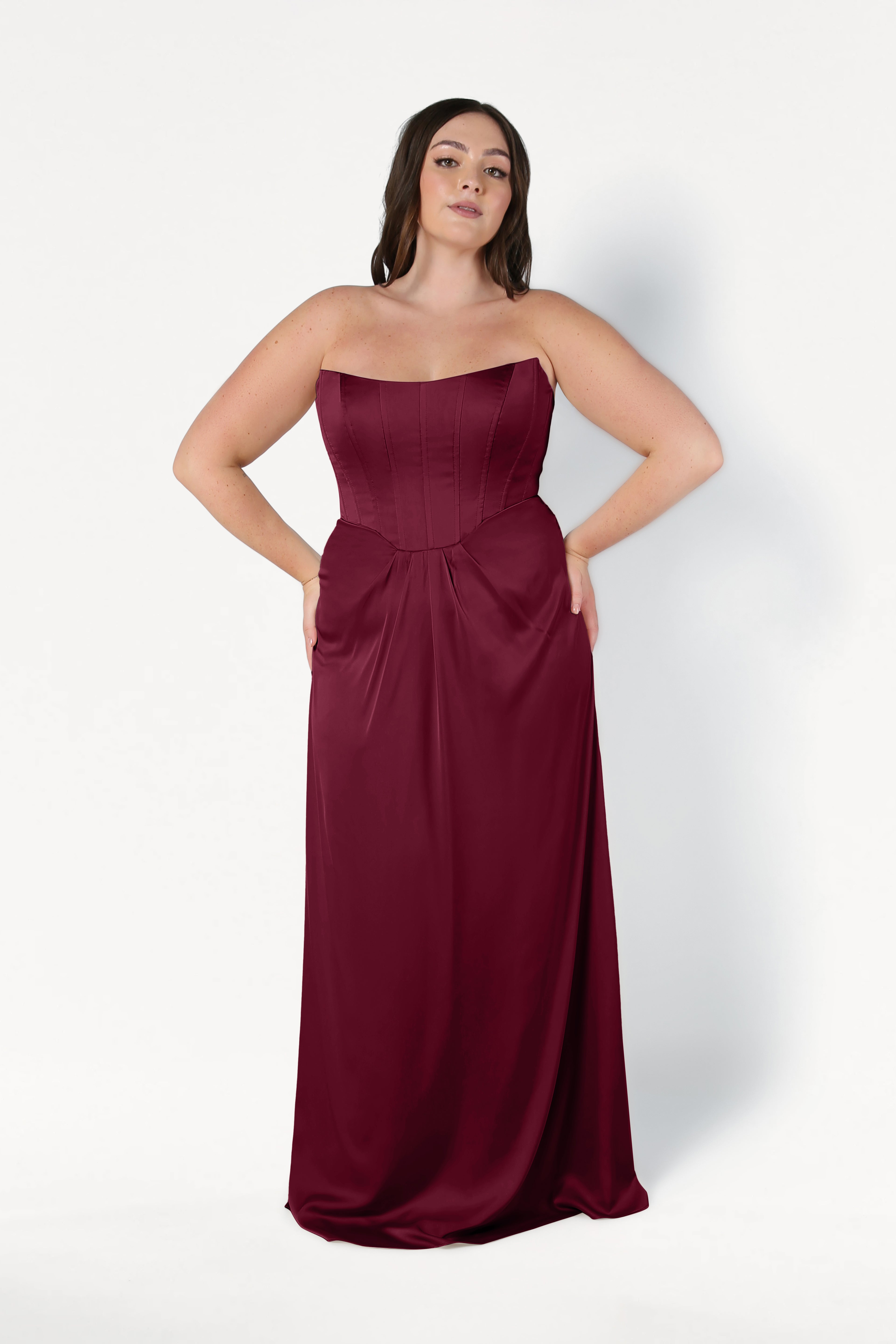 A woman wears the TH&TH Selena Bridesmaid Dress in Merlot Satin—a size-inclusive, strapless, floor-length gown with a fitted corset bodice and flowing skirt—standing against a white background with her hands in the dress pockets.