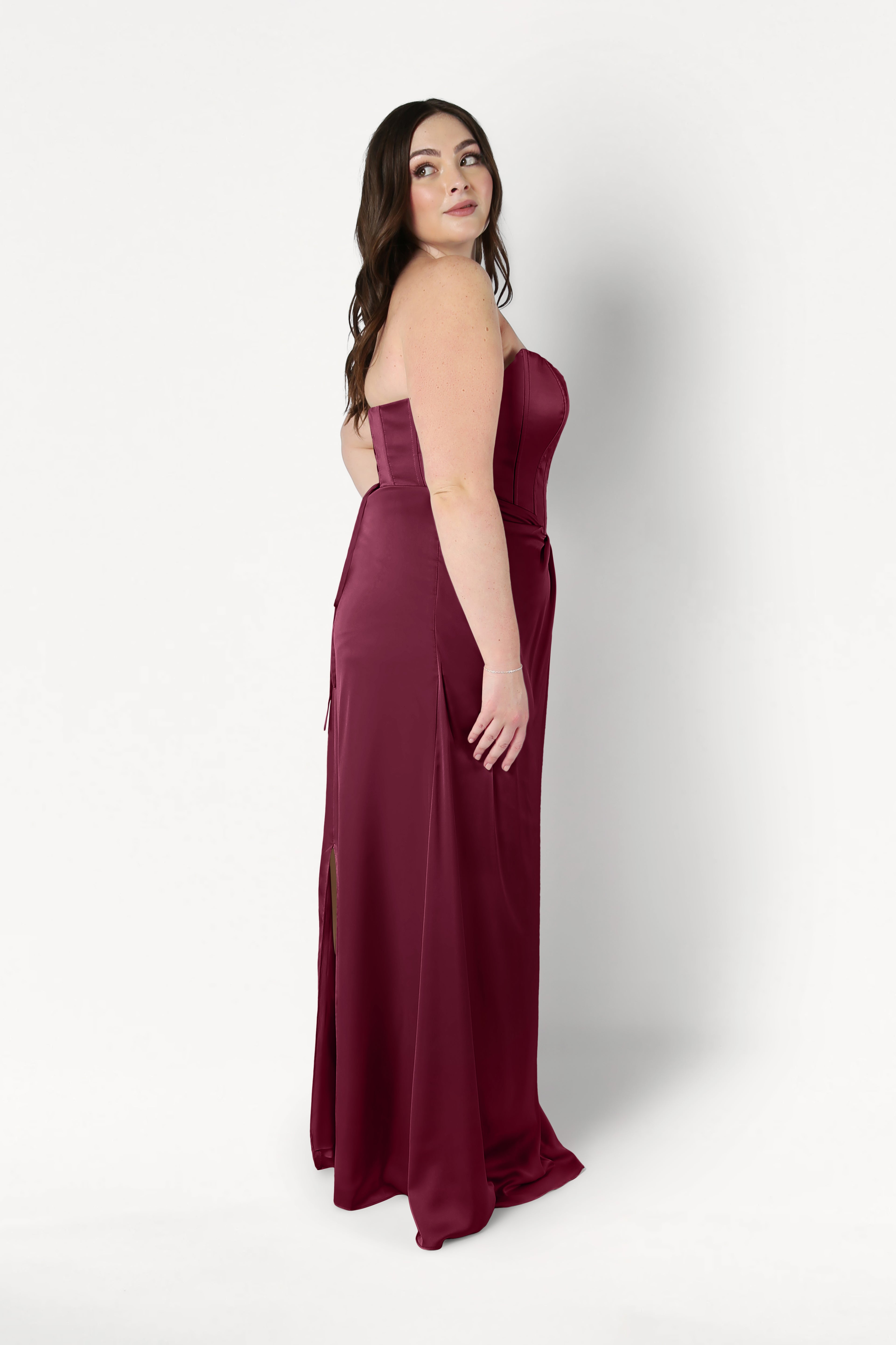 A woman with long brown hair models the TH&TH Selena Bridesmaid Dress in Merlot Satin—a size-inclusive, strapless gown with a corset bodice—standing sideways and looking over her shoulder against a white background.