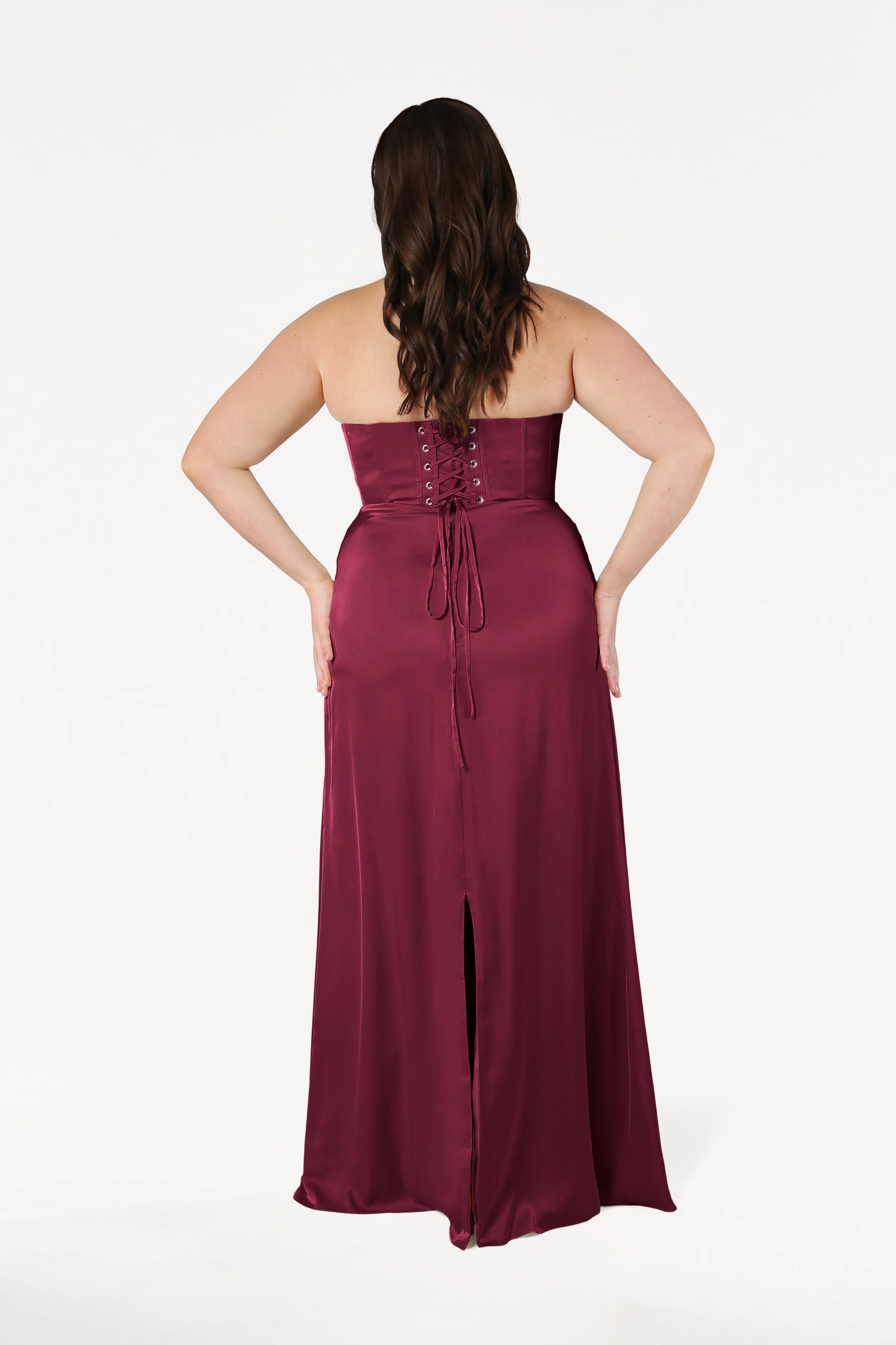 A woman with wavy brown hair models the size-inclusive Selena Bridesmaid Dress in Merlot Satin by TH&TH, featuring a strapless corset bodice with lace-up back and a long skirt with center slit, shown from behind against a white background.