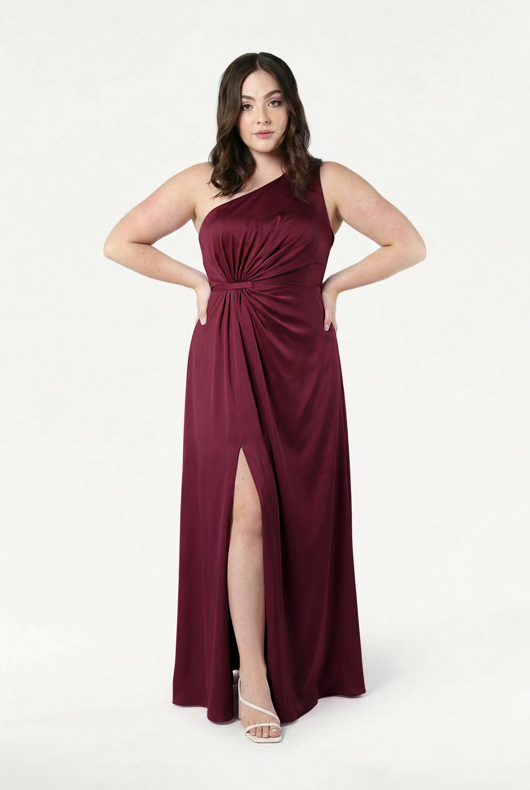 A woman stands against a white background wearing the Greta Bridesmaid Dress in Merlot Satin by TH&TH—a sleeveless, one-shoulder gown with a gathered knot detail and thigh-high slit. She pairs it with white heels and has mid-length brown hair.