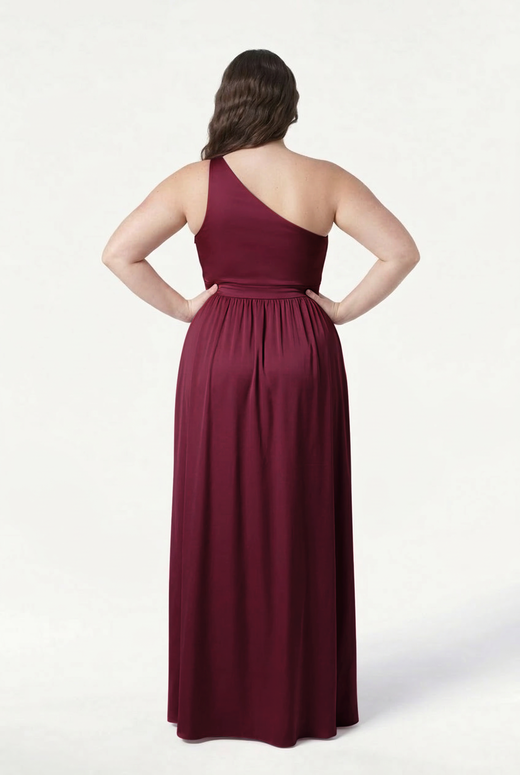 A woman stands with her hands on her hips, shown from the back in the TH&TH Greta Bridesmaid Dress—a floor-length, one-shoulder gown in Merlot satin—against a plain white background.