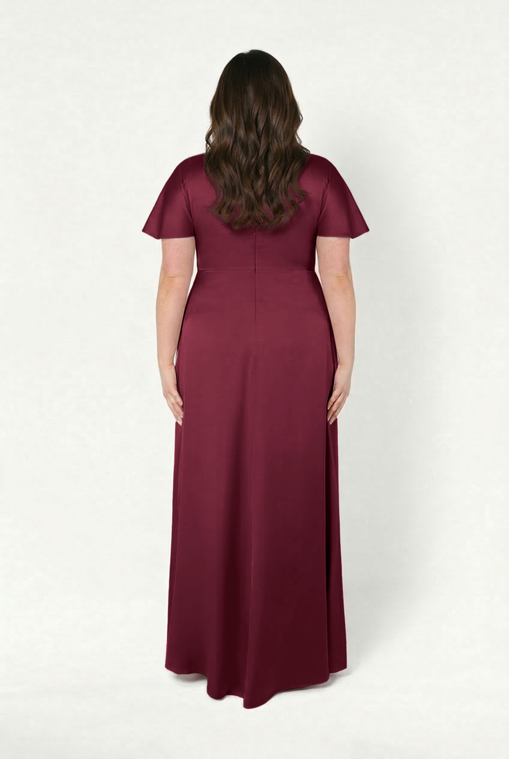 A woman with long brown hair stands facing away, wearing the TH&TH Camilla Bridesmaid Dress in Merlot Satin, a modern floor-length gown, against a plain white background.