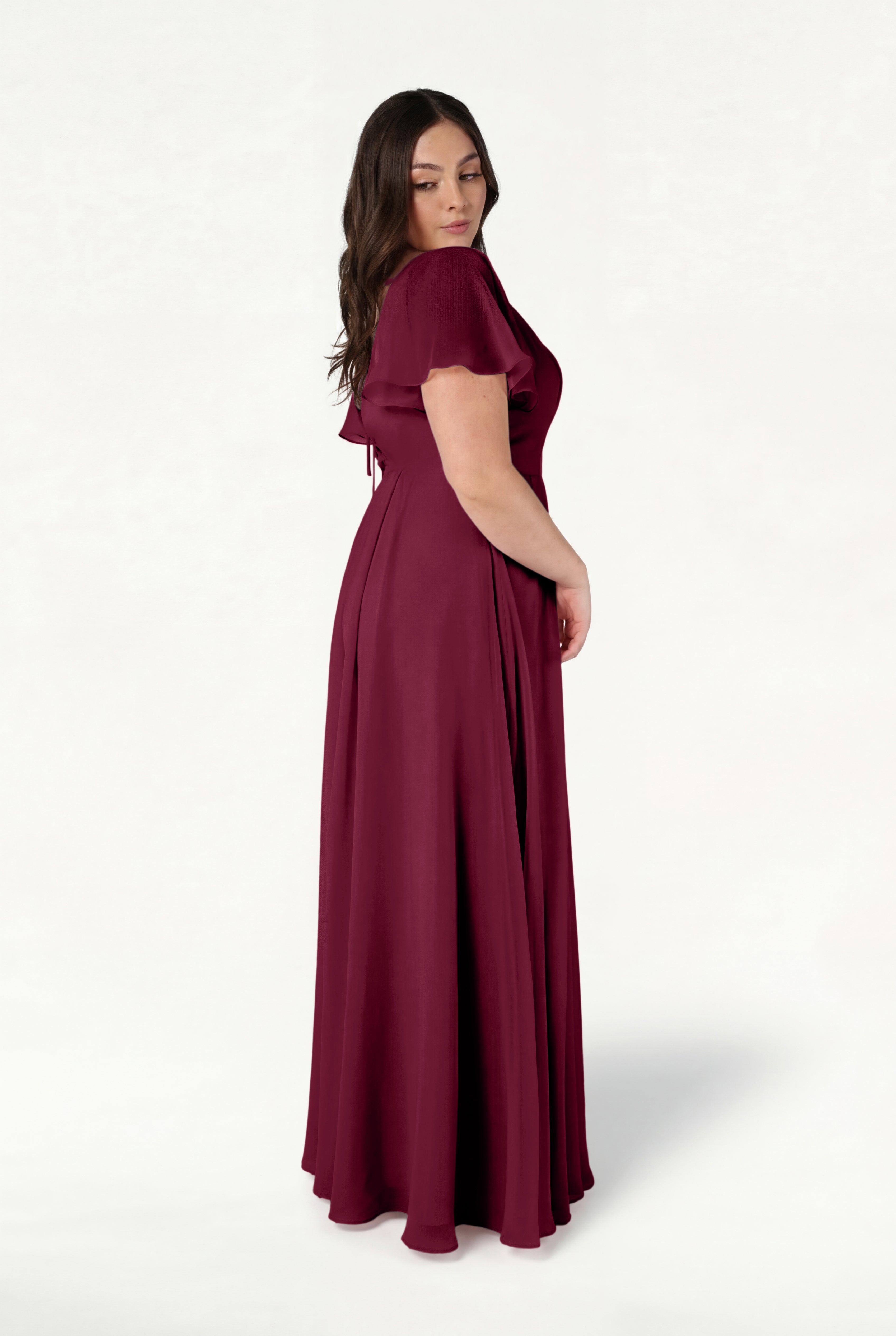 A woman models the TH&TH Phoebe Bridesmaid Dress in Merlot Chiffon, a floor-length, short-sleeved gown. She stands sideways against a plain white background, glancing over her shoulder with her long brown hair down.