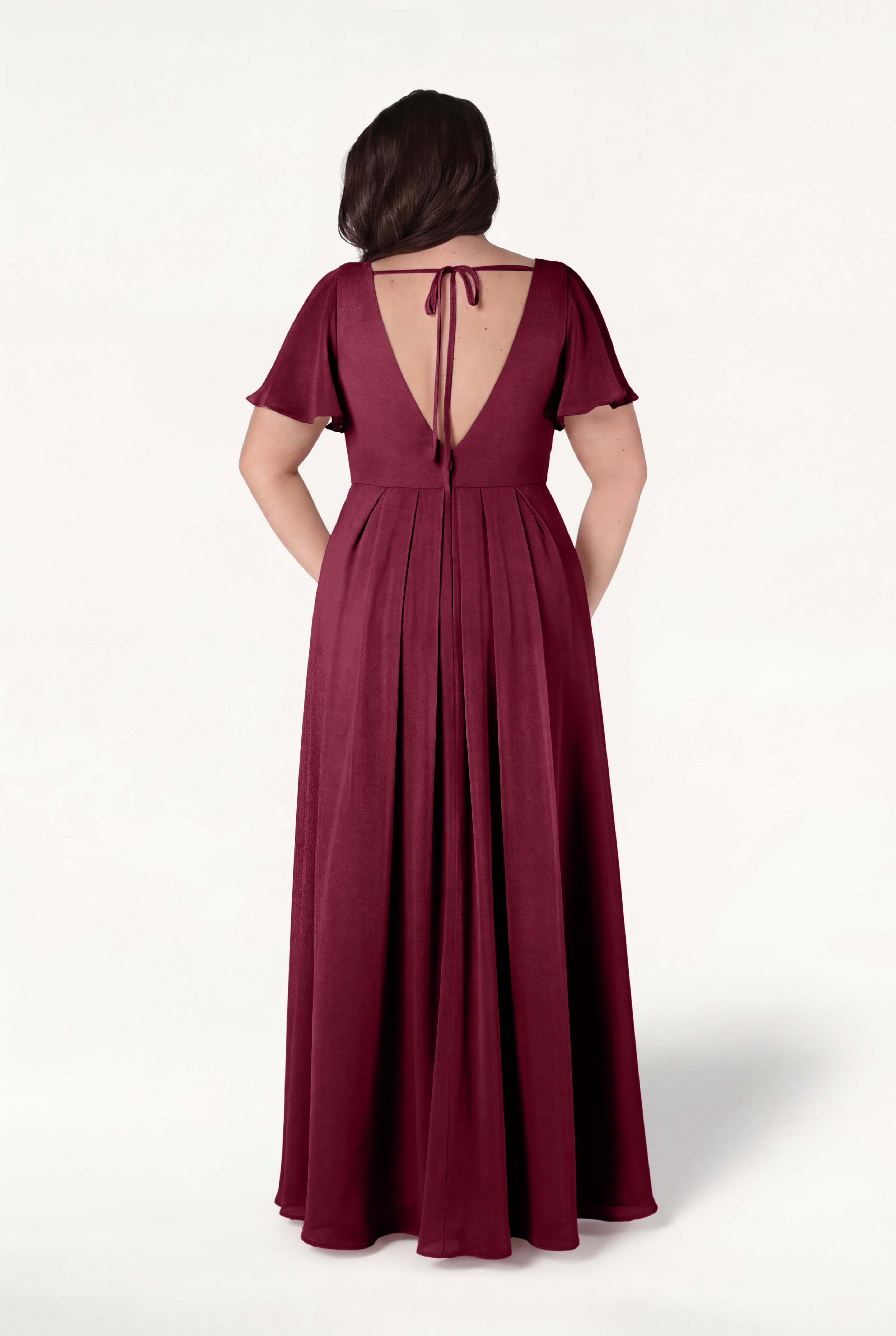 A woman faces away, wearing the TH&TH Phoebe Bridesmaid Dress in Merlot Chiffon—a size-inclusive, floor-length gown with short flutter sleeves, a low tie-back, and flowing fabric—set against a plain light background.