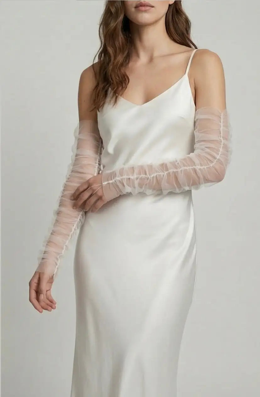 A woman poses against a plain background wearing TH&TH's Millie Ruched Tulle Opera Sleeves in ivory over a white satin slip dress with spaghetti straps. Her wavy hair frames her face as she stands with one arm bent.