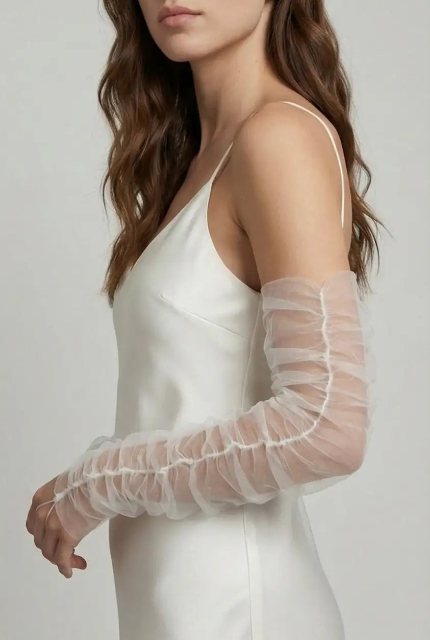 A woman with wavy brown hair stands sideways against a plain light background, wearing TH&TH’s Millie Ruched Tulle Opera Sleeves in Ivory over a silky white dress for a romantic touch; her face is partially out of frame.