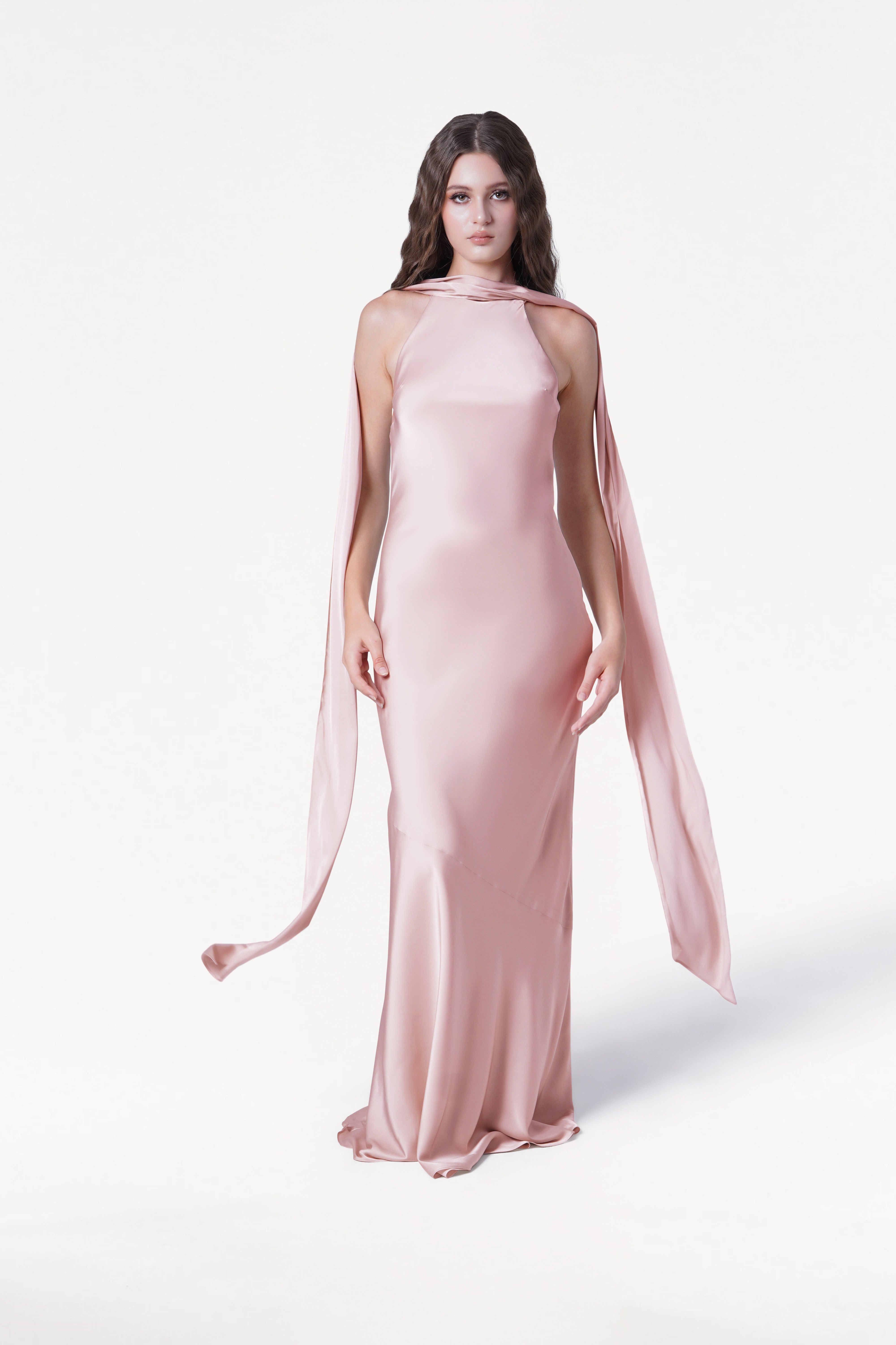 A woman with long, wavy brown hair stands against a plain white background, wearing the TH&TH Mira Bridesmaid Dress in blush pink satin, featuring a high neckline and flowing scarf-like fabric draped from the shoulders.