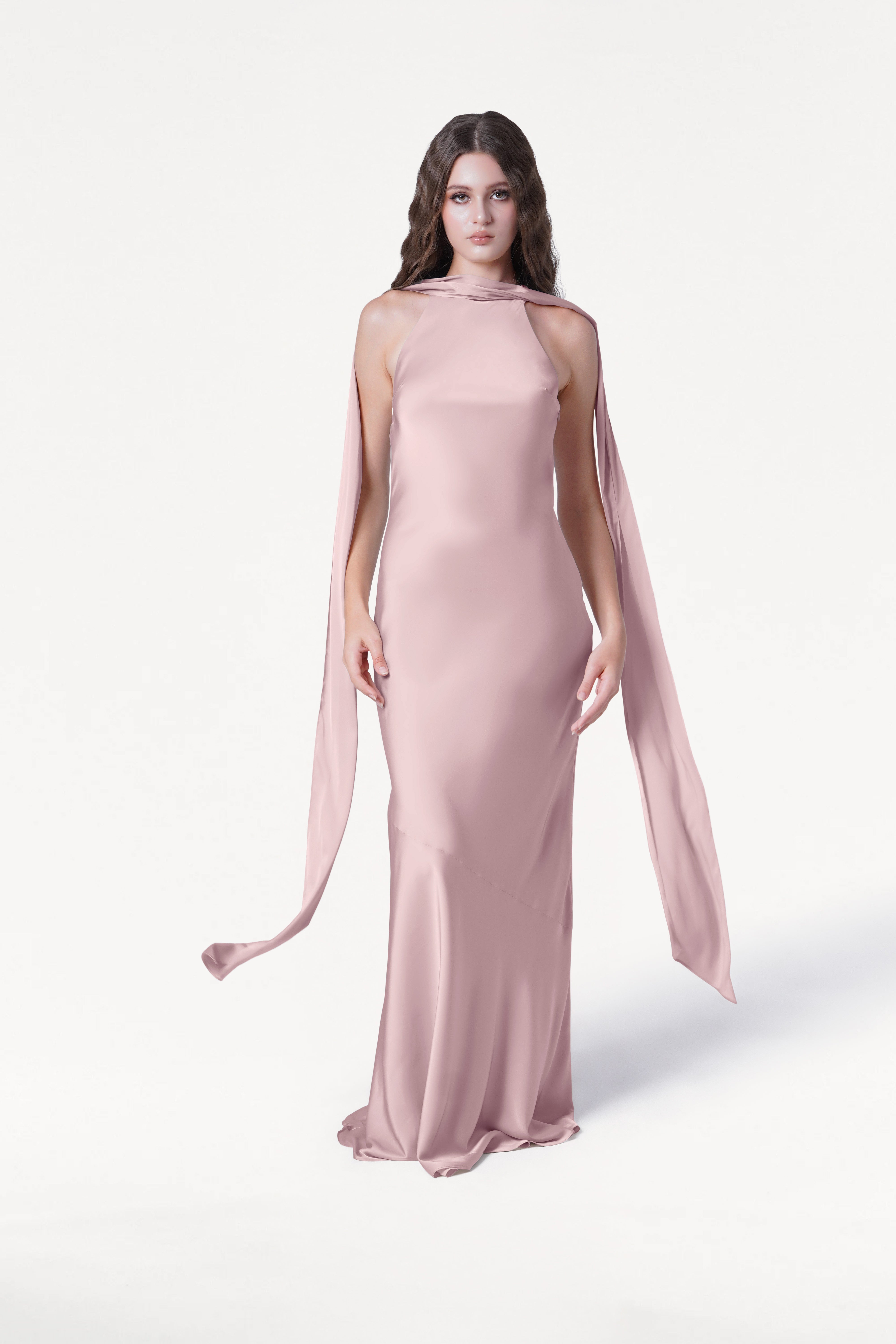 A woman with long, wavy brown hair and a neutral expression models the TH&TH Mira Bridesmaid Dress in Smoked Blush Satin, featuring long shoulder sashes and a high neckline, against a white background.