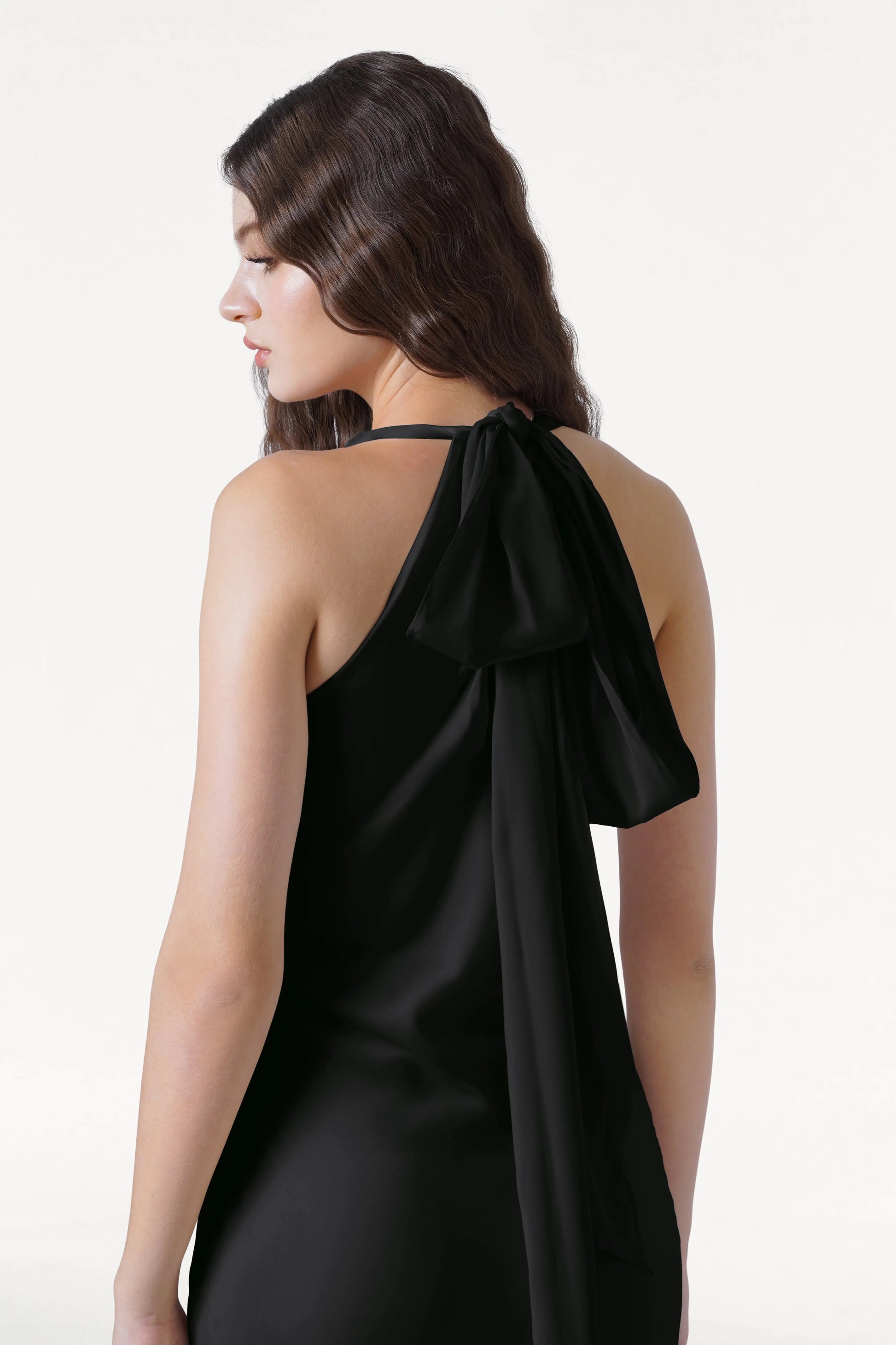 A woman with long wavy brown hair wears the TH&TH Mira Bridesmaid Dress in Black Satin, featuring a black halterneck design with a large bow at the back, standing against a plain white background.