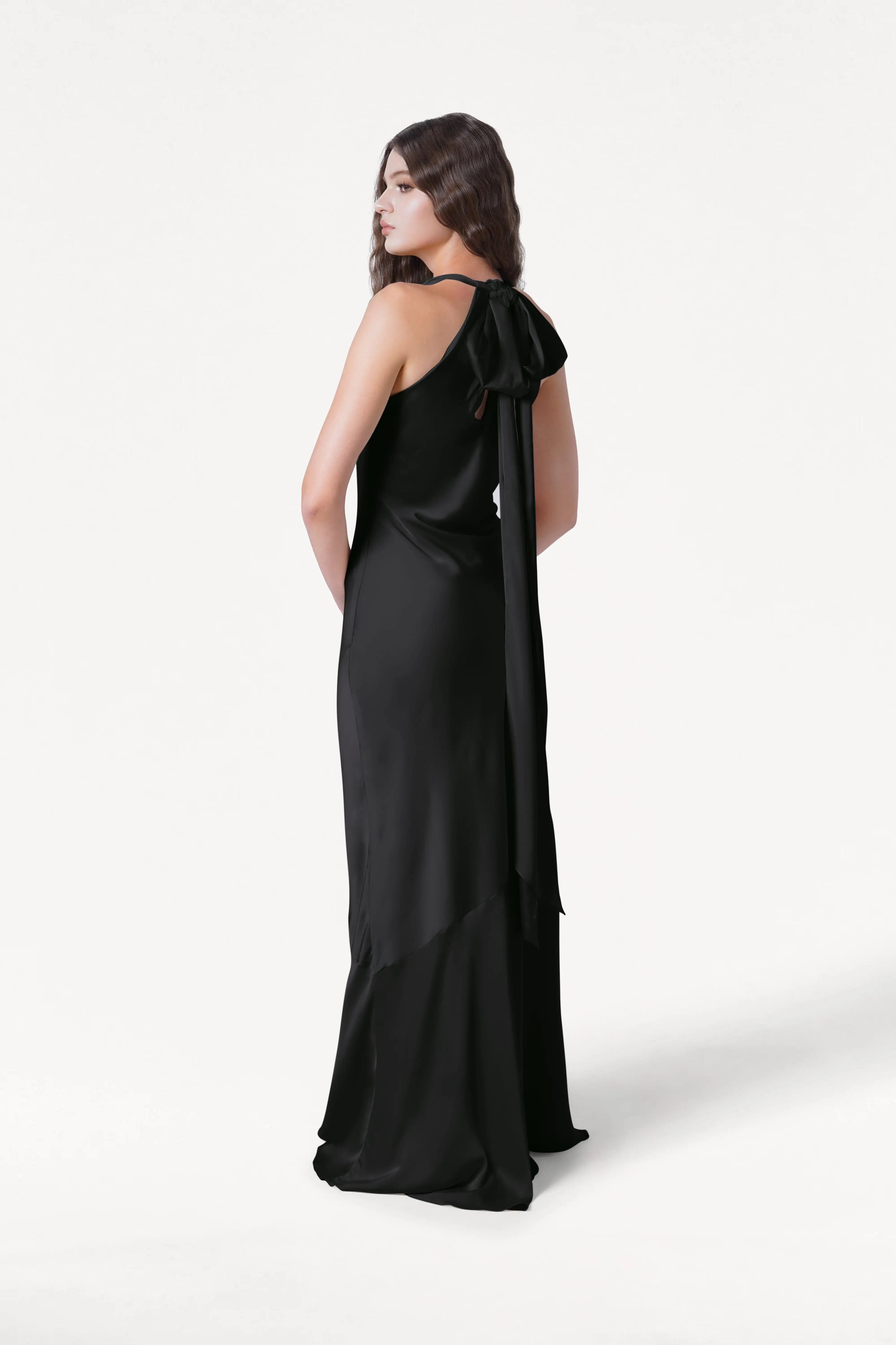 A woman with long brown hair models the TH&TH Mira Bridesmaid Dress in Black Satin, a sleeveless, floor-length gown with an asymmetrical hem and a large shoulder bow, standing against a plain white background.