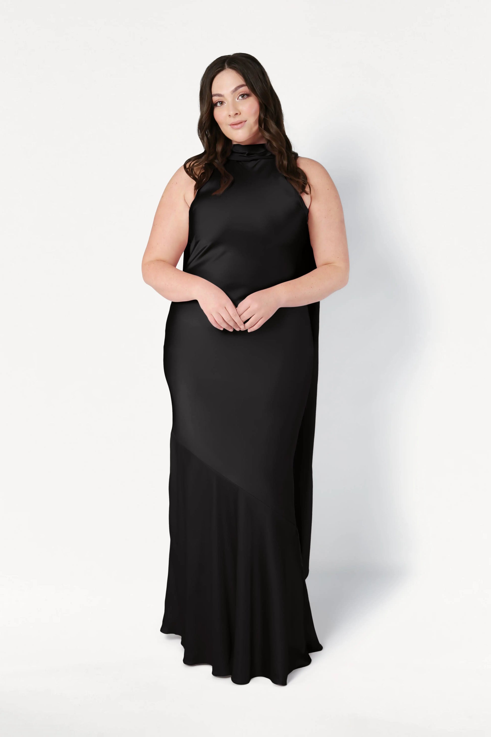 A woman with long brown hair wears the Mira Bridesmaid Dress in Black Satin by TH&TH. She stands against a plain white background, facing forward with her hands gently clasped in front of her.