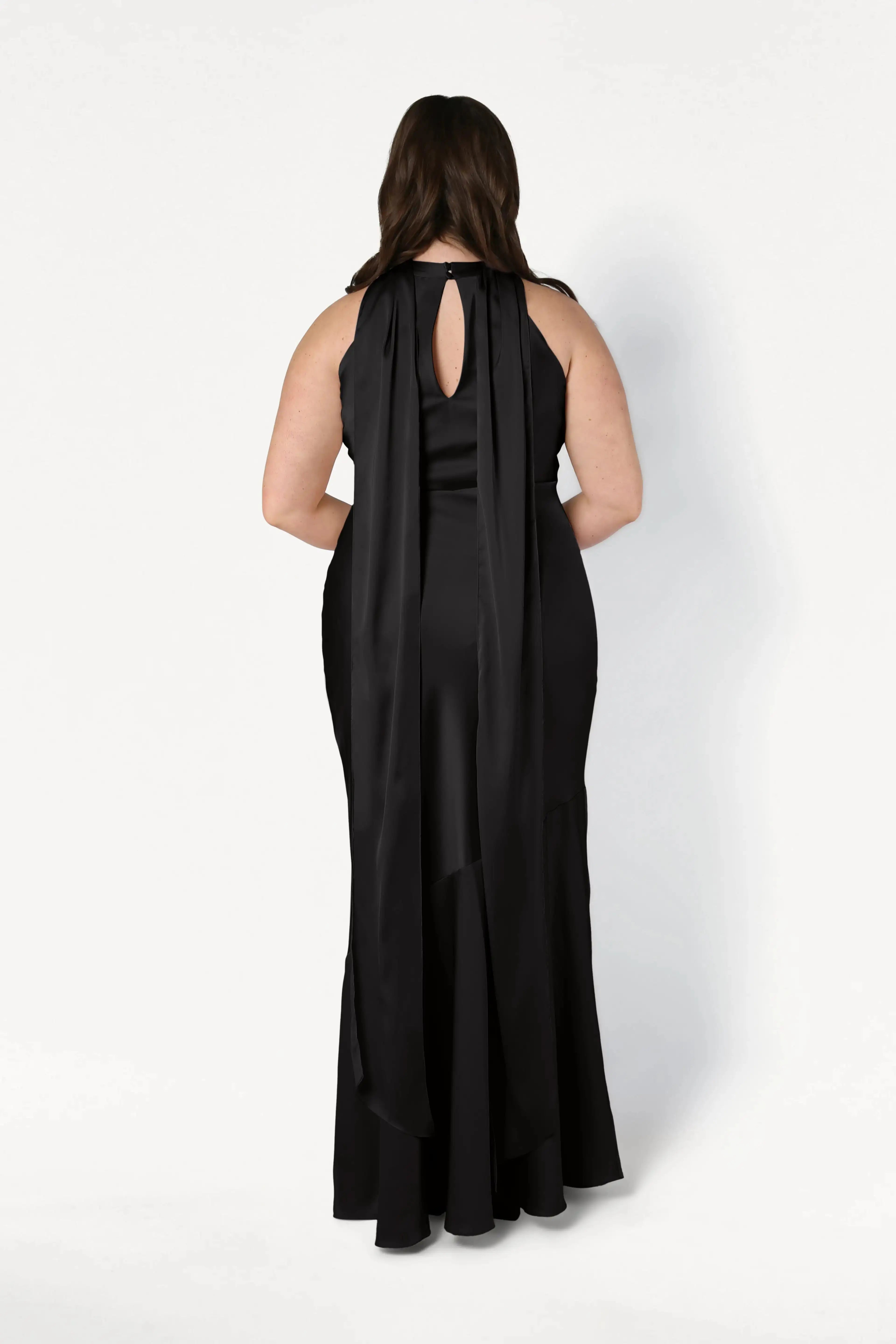 A woman wears the TH&TH Mira Bridesmaid Dress in black satin, showing its long, sleeveless design with a keyhole back and flowing shoulder panels as she poses facing away against a plain white background.