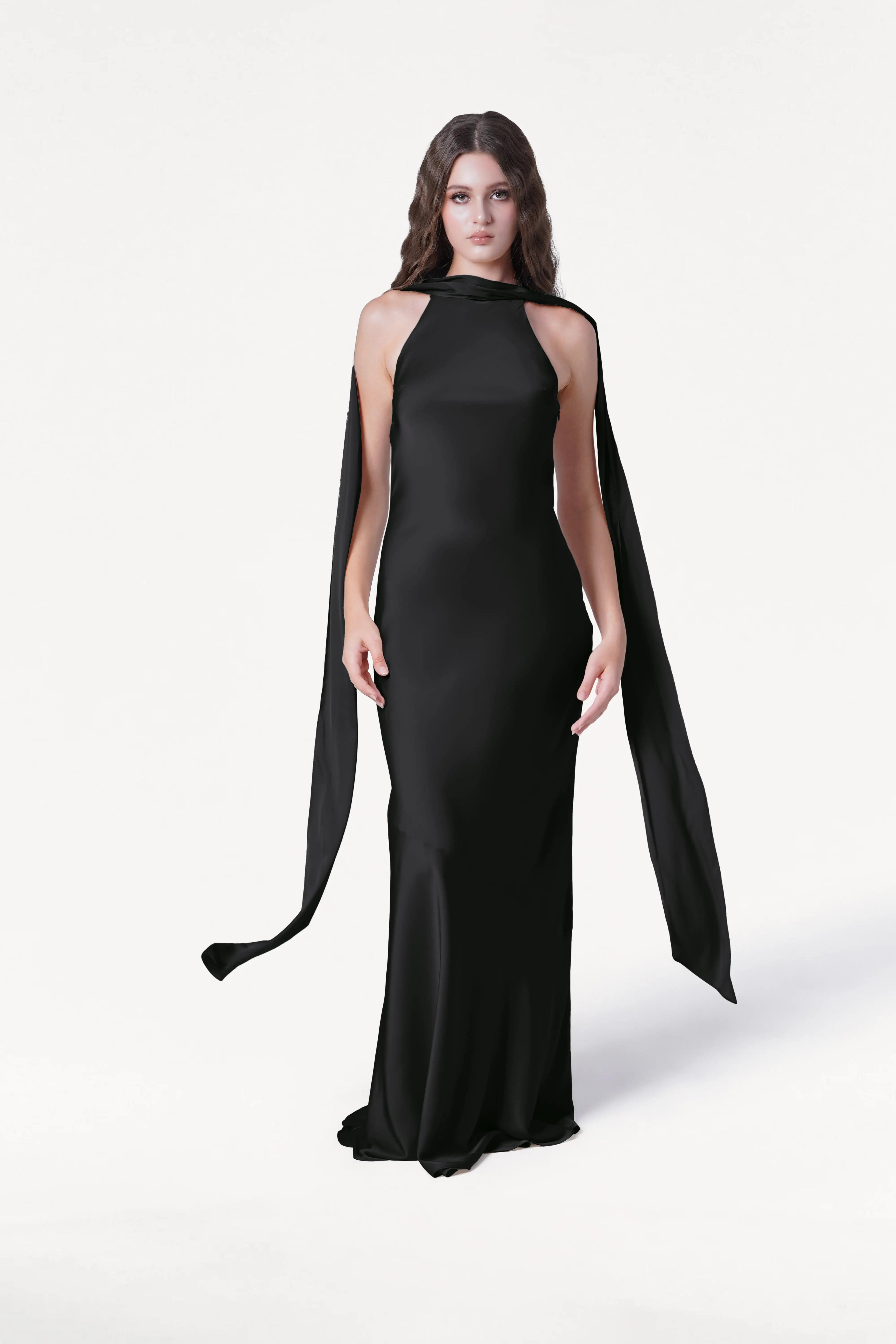 A woman with long wavy hair wears the Mira Bridesmaid Dress in Black Satin by TH&TH, featuring a floor-length, bias-cut silhouette with flowing cape-like panels, standing against a plain white background.