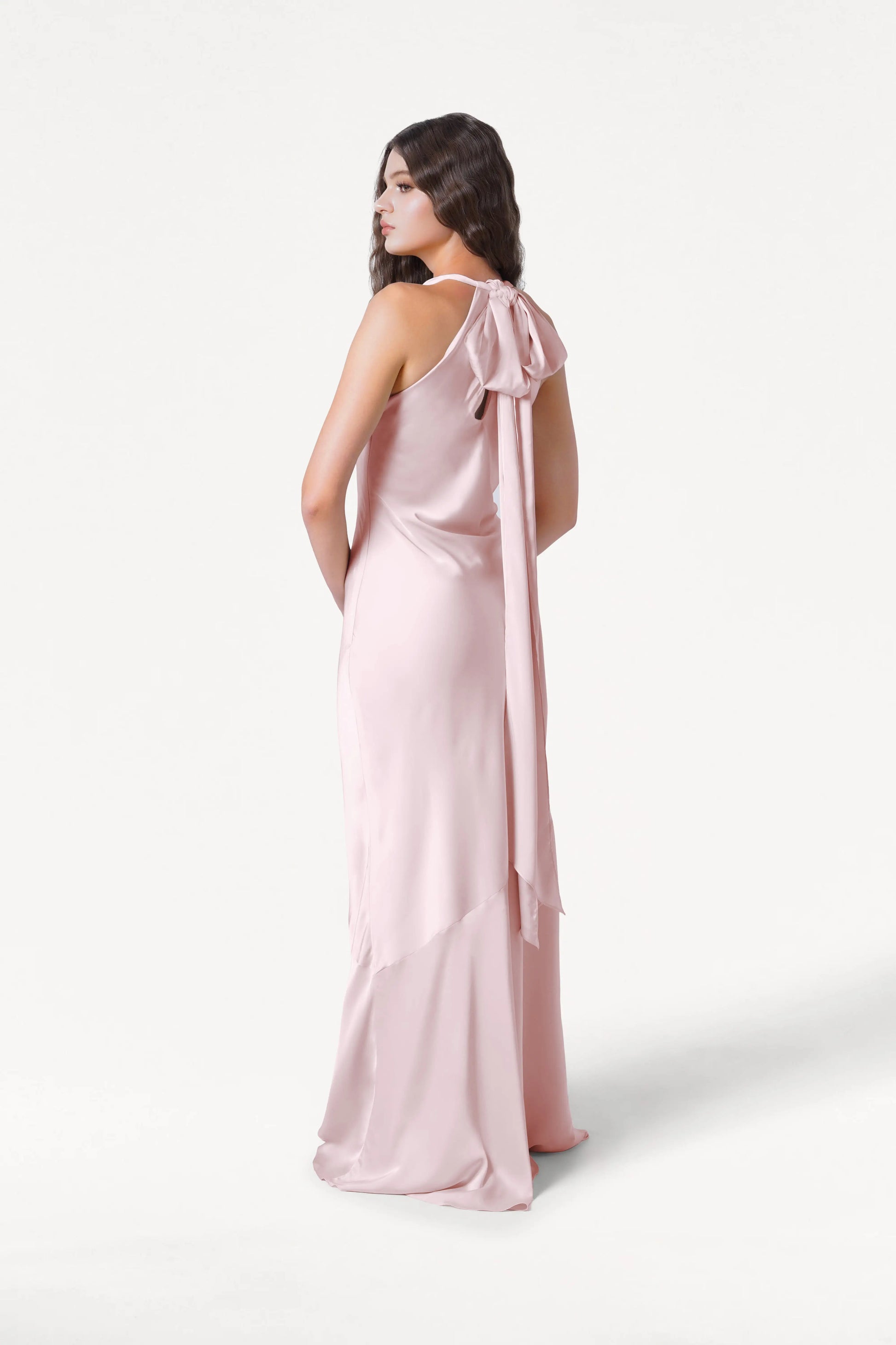 A woman models the TH&TH Mira Bridesmaid Dress in Blush Pink Satin, featuring a tie-back detail and floor-length smooth satin, as she stands facing away with her head turned to the side against a white background.