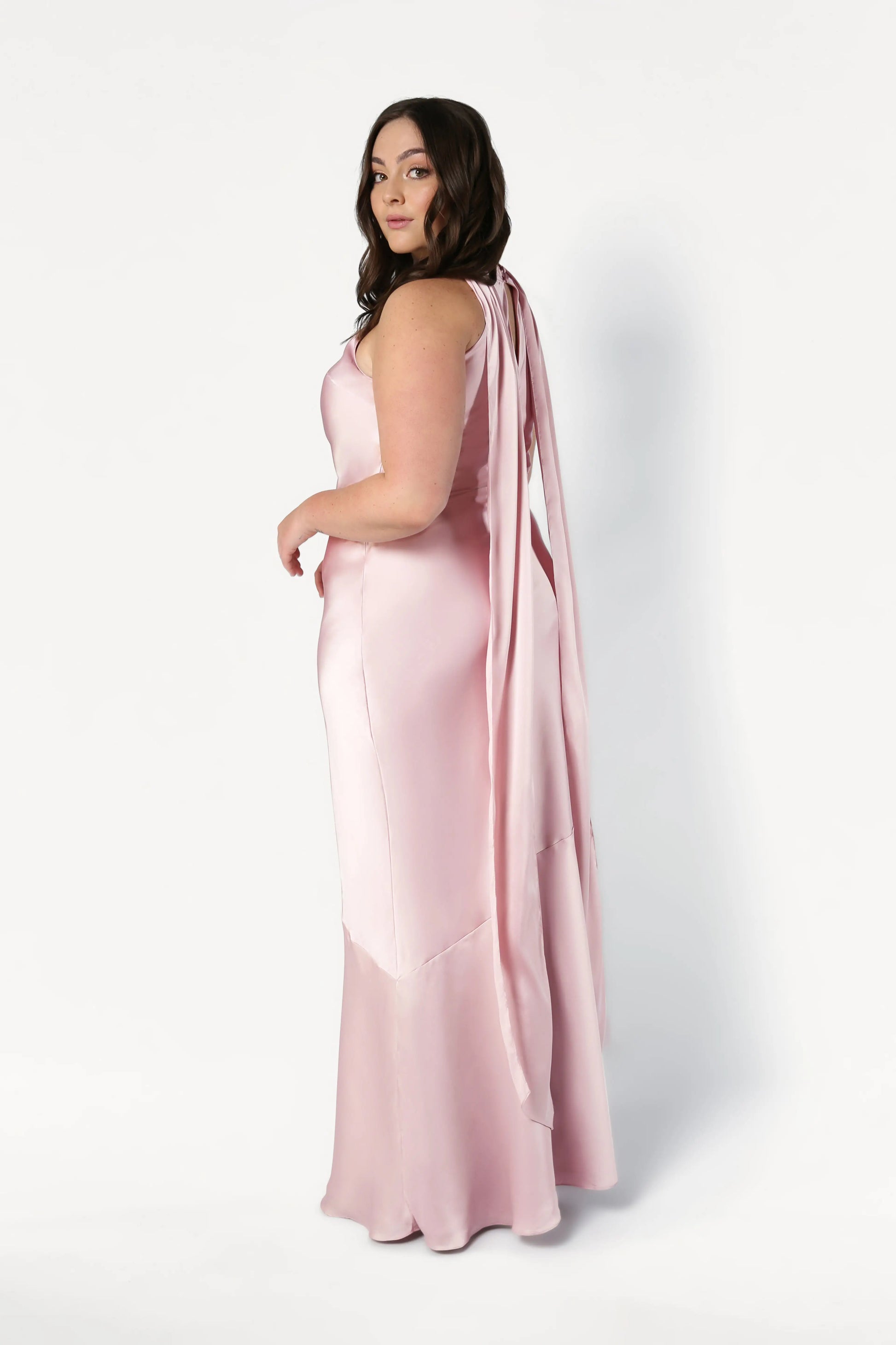 A woman with long brown hair poses sideways against a white background wearing the TH&TH Mira Bridesmaid Dress in blush pink satin, featuring a floor-length silhouette and long ribbon details.