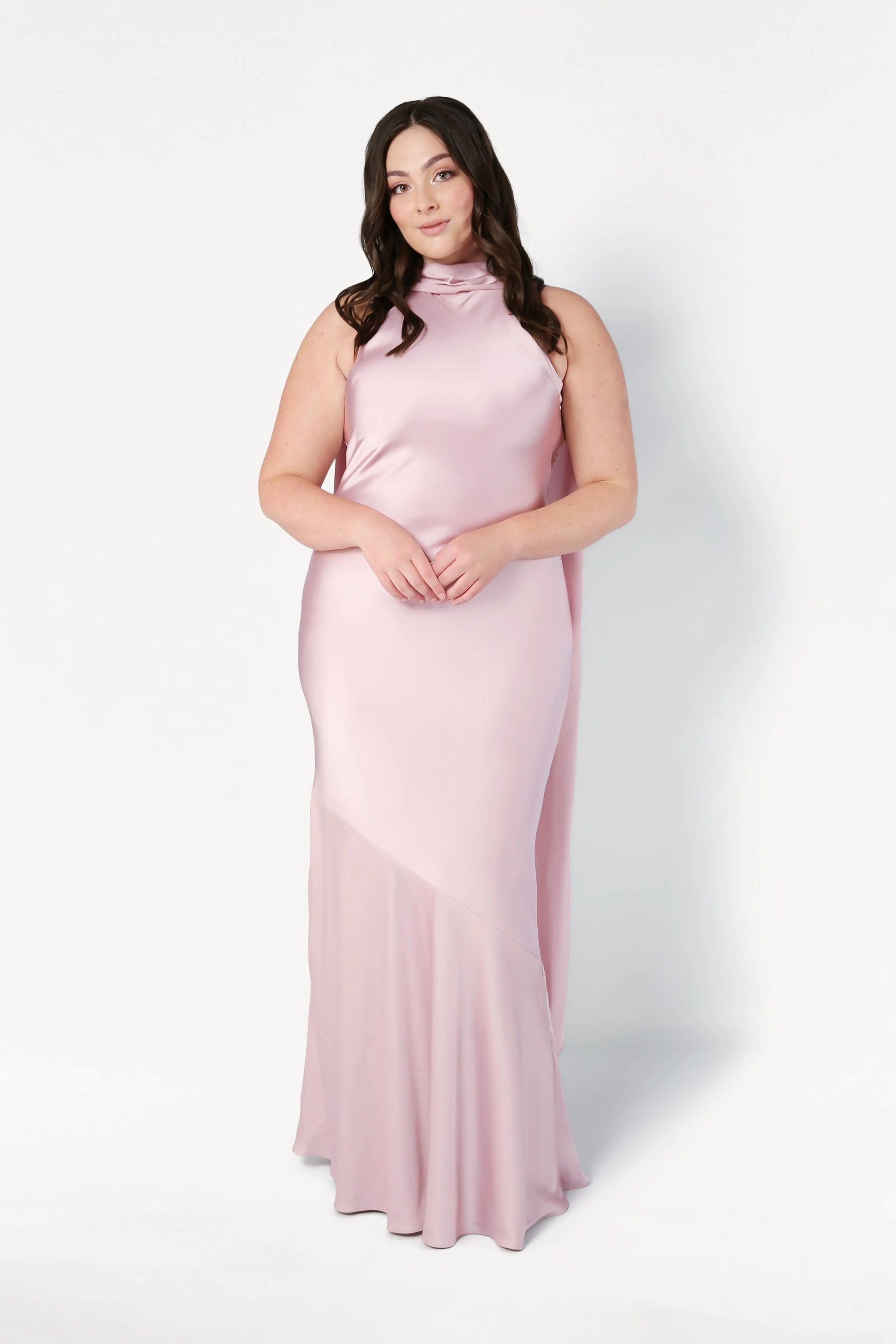 A woman with long dark hair wears the TH&TH Mira Bridesmaid Dress in blush pink satin, featuring a sleeveless design and high neckline, standing against a plain white background.
