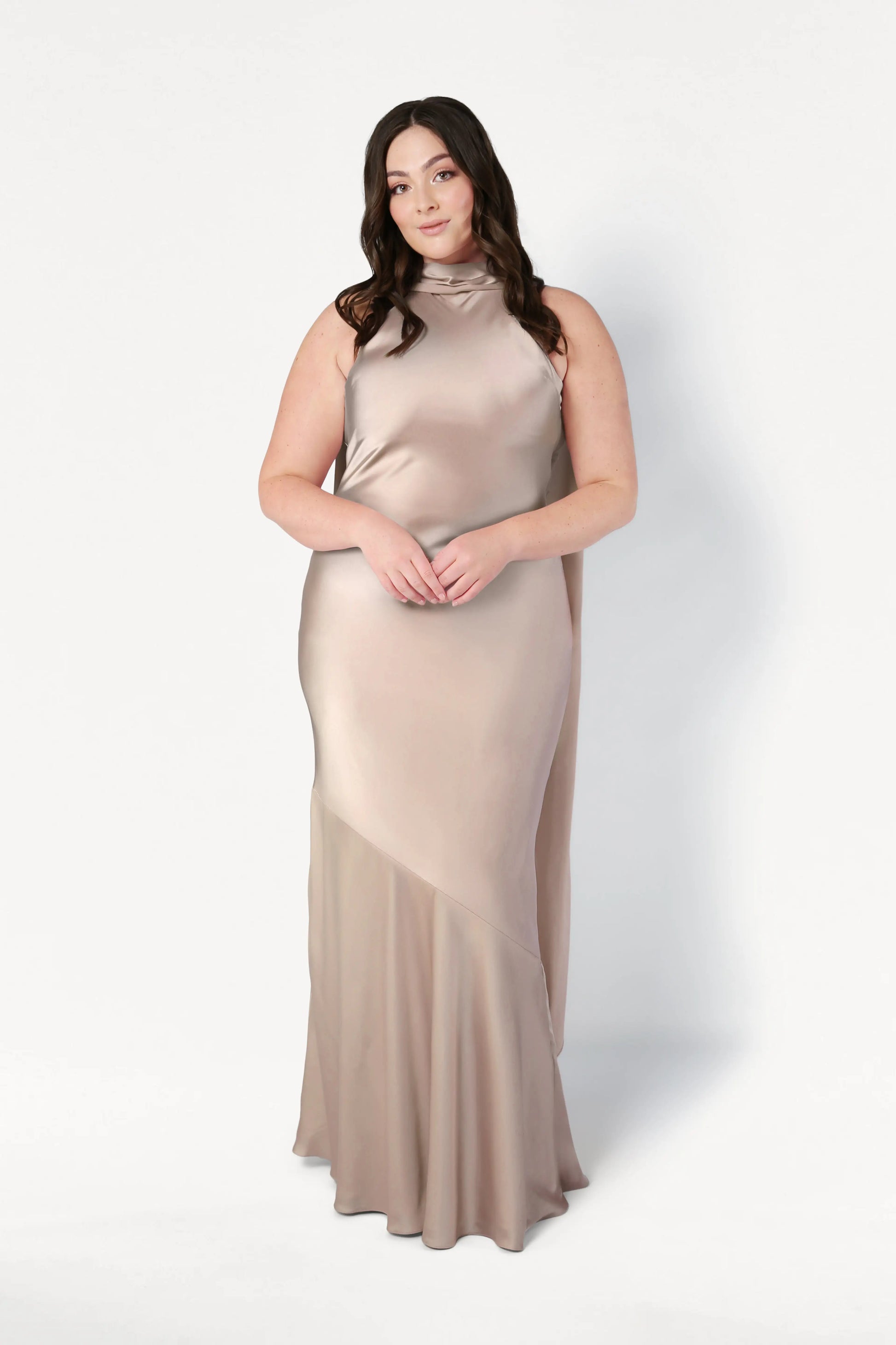 A woman with long dark hair wears the TH&TH Mira Bridesmaid Dress in Champagne Sky Satin. She stands against a plain white background, gazing at the camera with a gentle expression.
