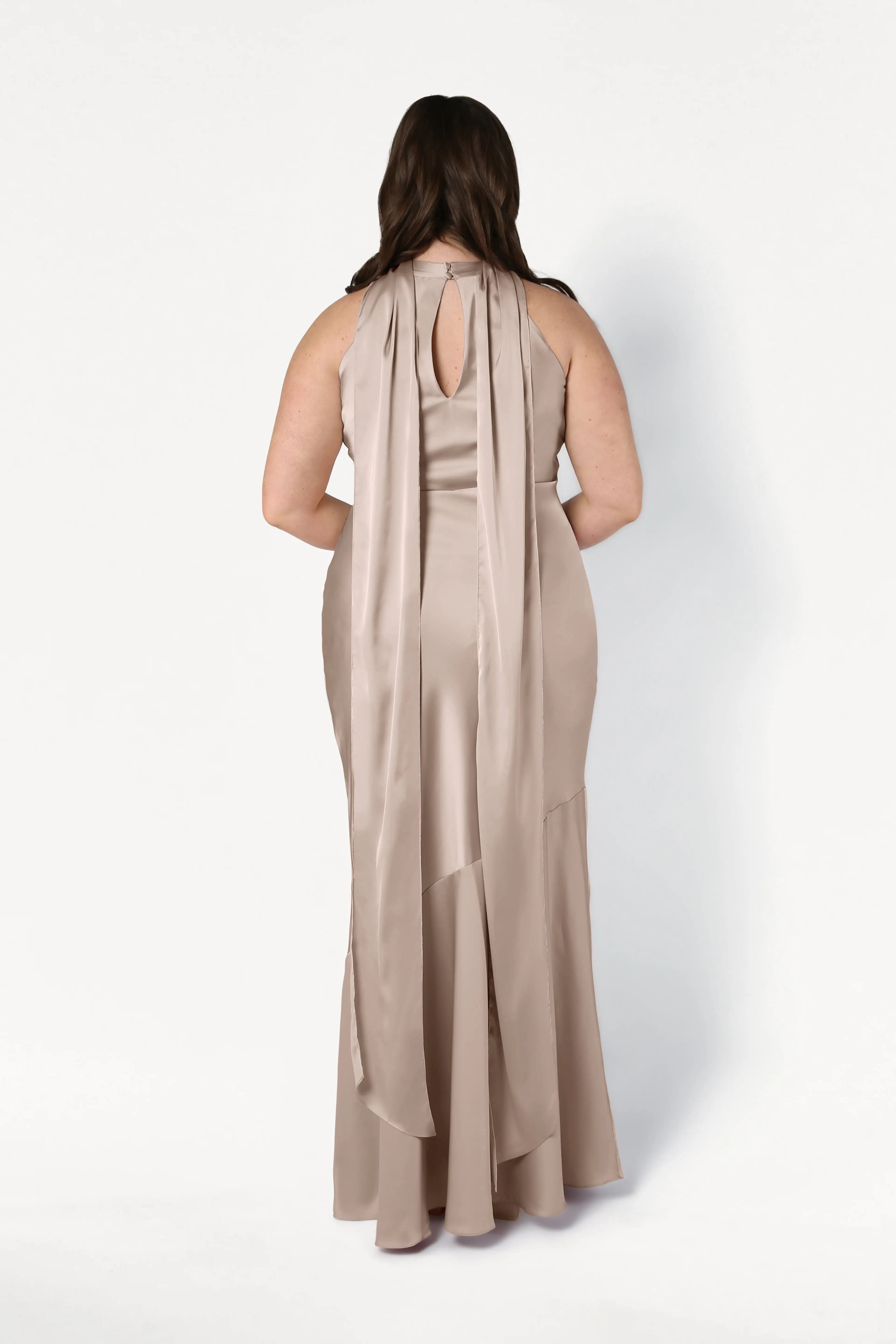 A woman faces away, wearing the Mira Bridesmaid Dress in Champagne Sky Satin by TH&TH. The dress features a keyhole back with two flowing ribbons from the shoulders against a plain white background.
