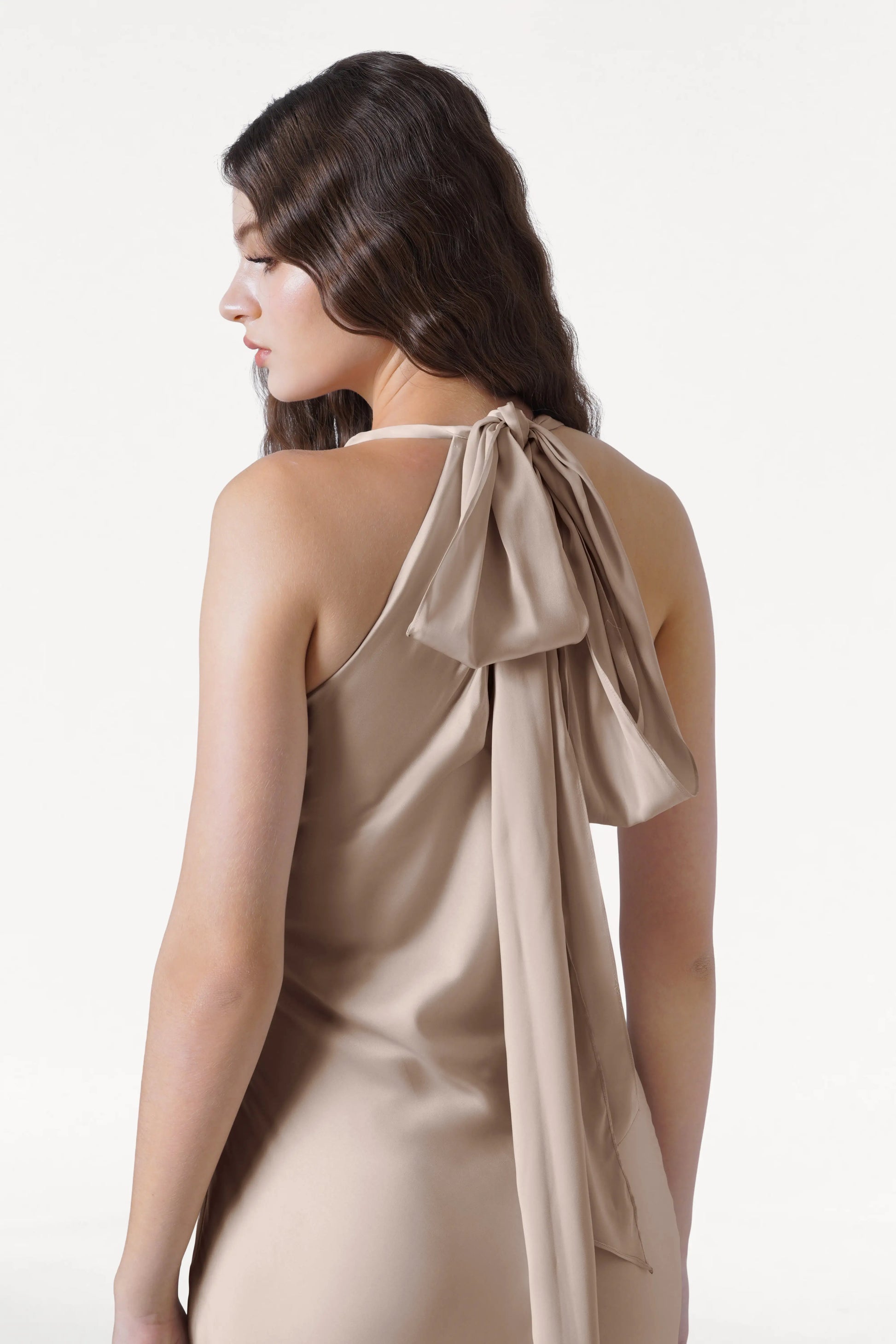 A woman with long, wavy brown hair models the TH&TH Mira Bridesmaid Dress in Champagne Sky Satin, featuring a one-shoulder design with a large bow and tail at the back. She stands against a plain white background, looking to the side.