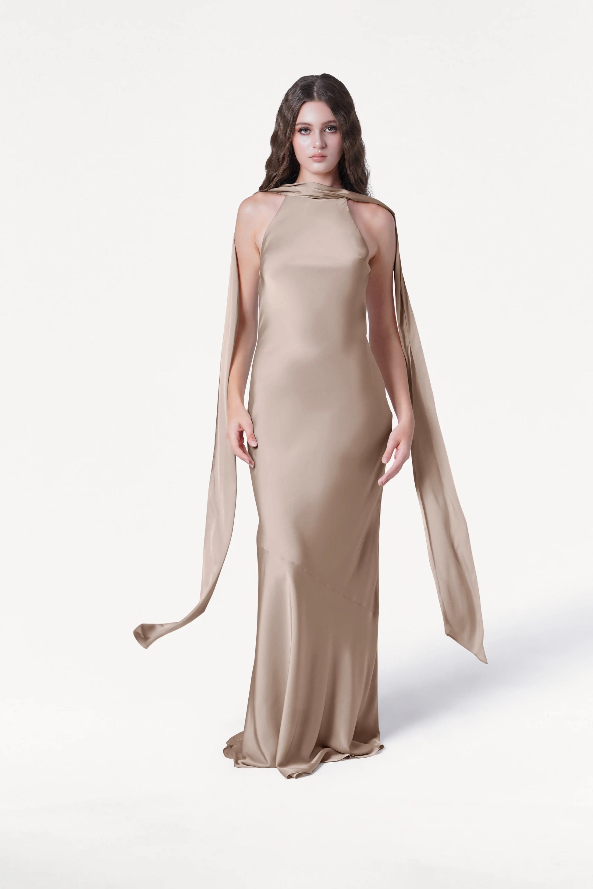 A woman with long, wavy brown hair and a neutral expression wears the TH&TH Mira Bridesmaid Dress in Champagne Sky Satin, featuring a floor-length silhouette, high neckline, and long, flowing side panels against a white background.