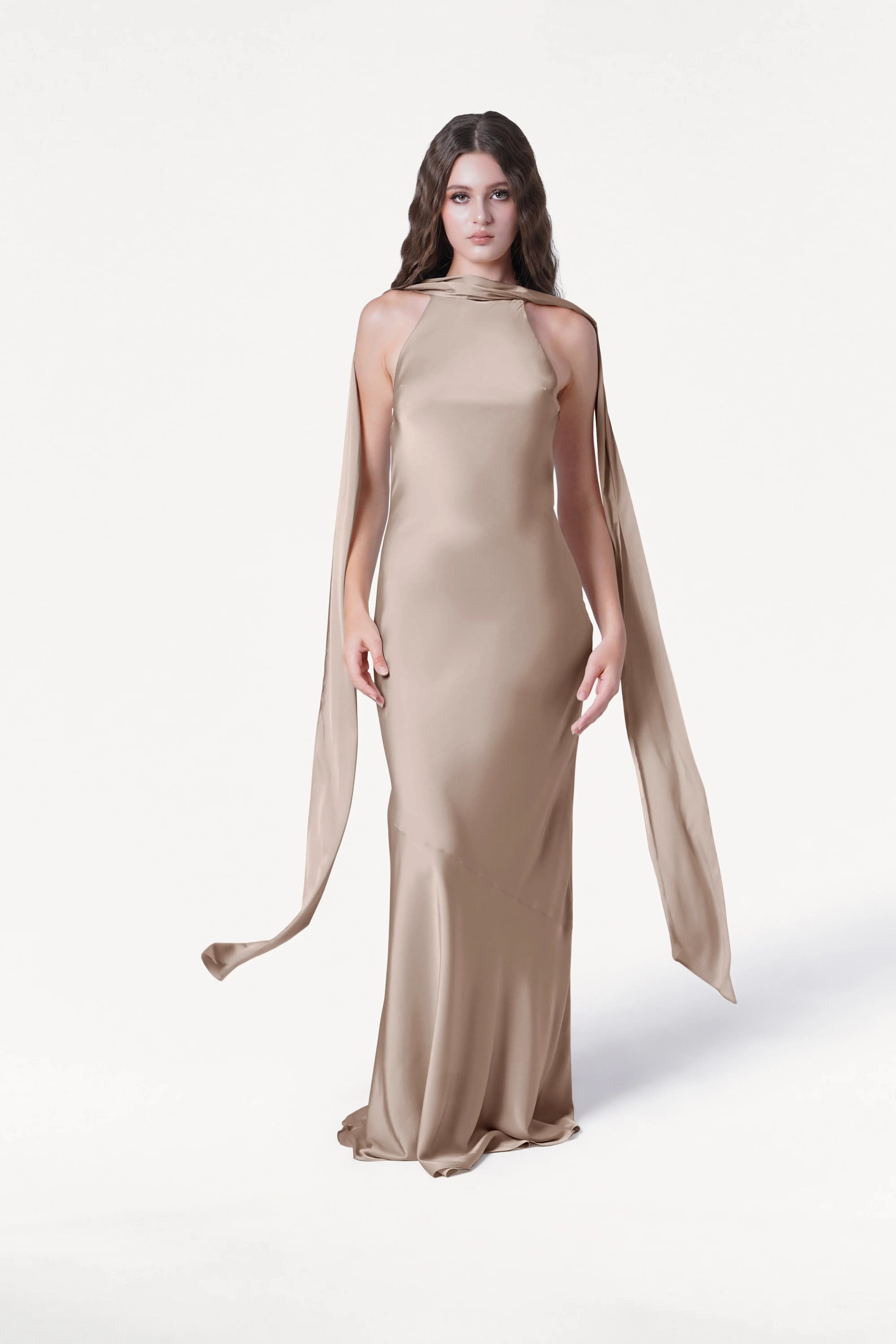A woman with long, wavy brown hair and a neutral expression wears the TH&TH Mira Bridesmaid Dress in Champagne Sky Satin, featuring a floor-length silhouette, high neckline, and long, flowing side panels against a white background.